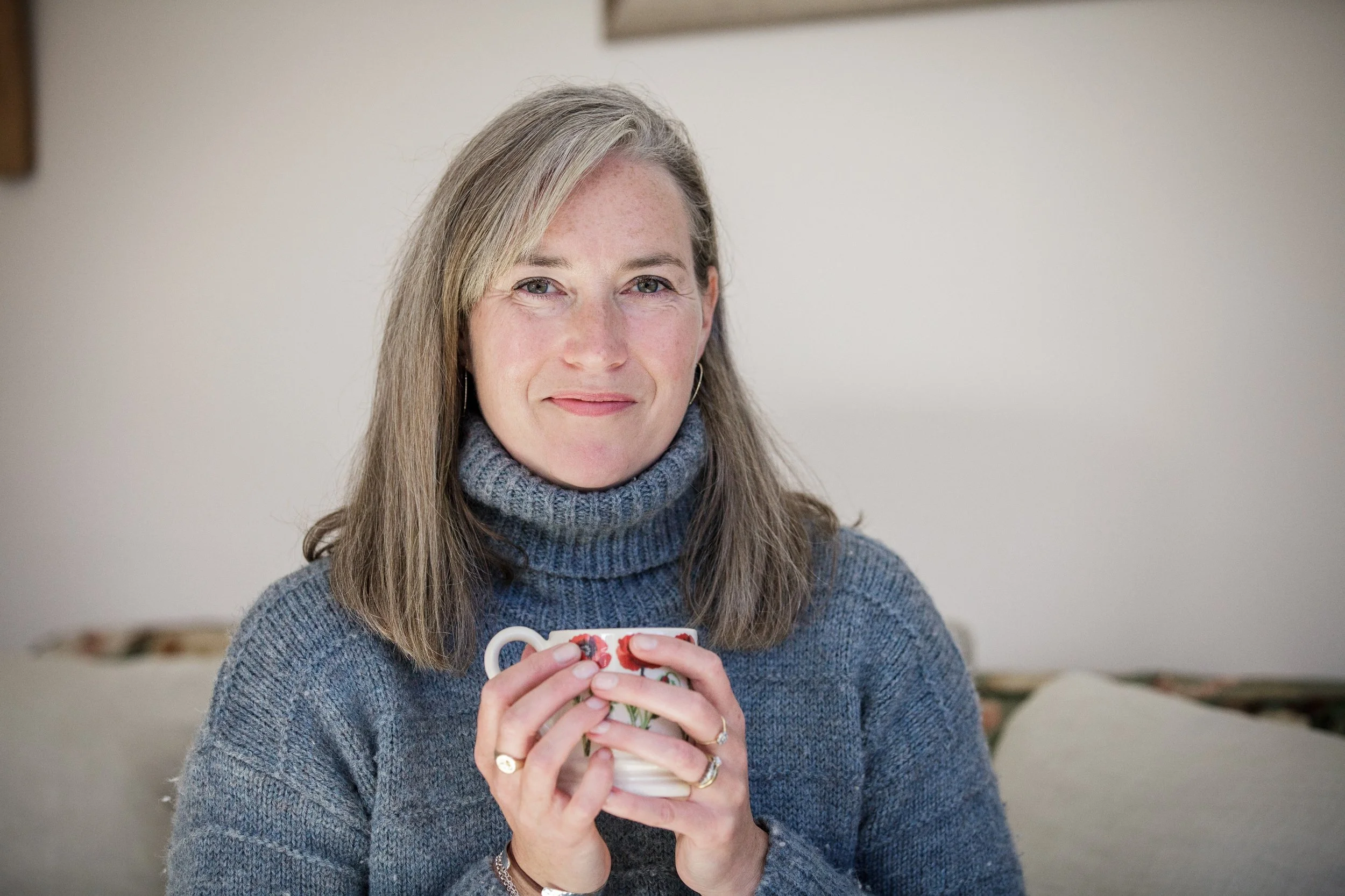 A woman with gray hair wearing a blue turtleneck sweater holding a patterned ceramic mug.