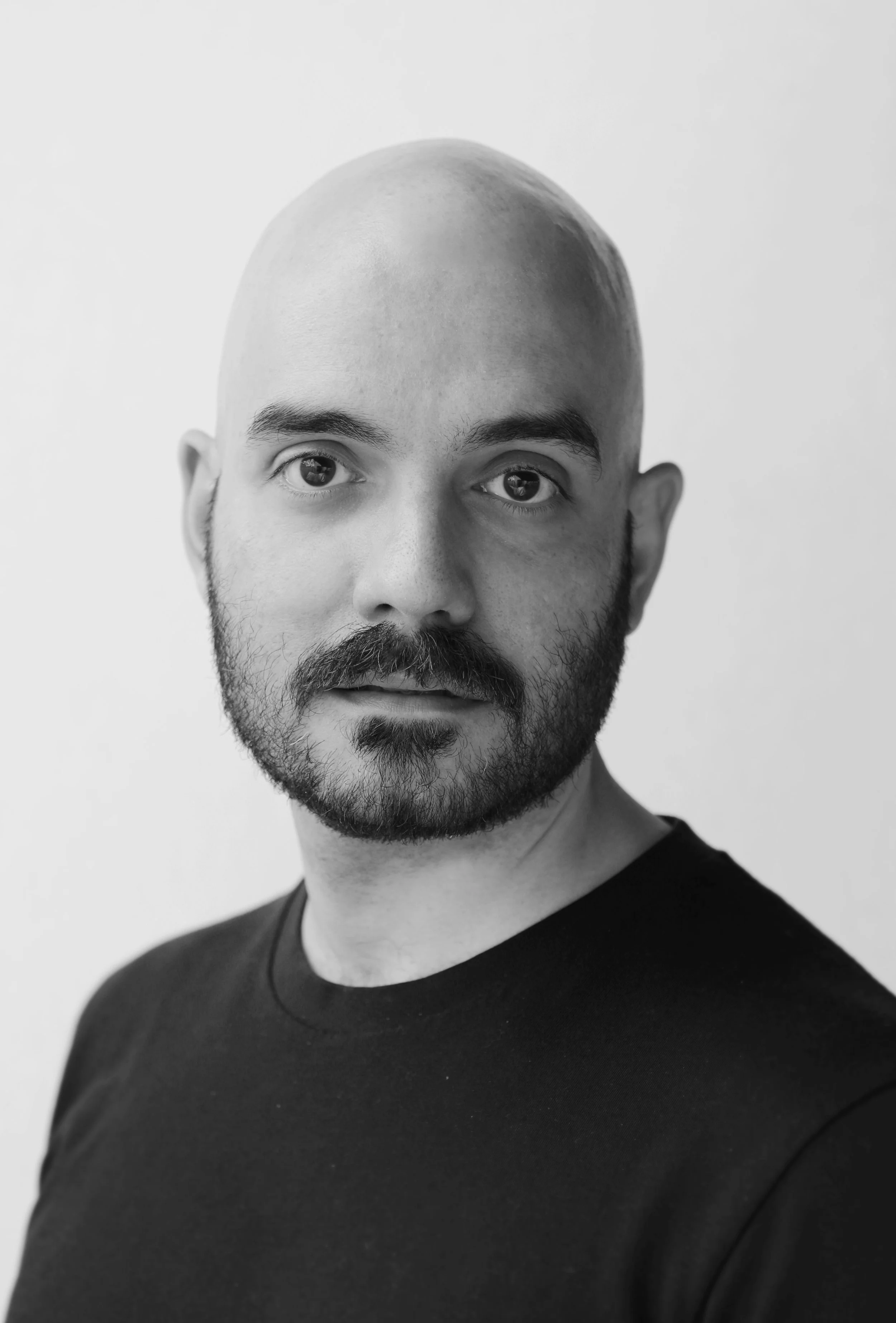 Black and white portrait of a man with no hair, a full beard, wearing a dark shirt, looking directly at the camera against a plain light background.