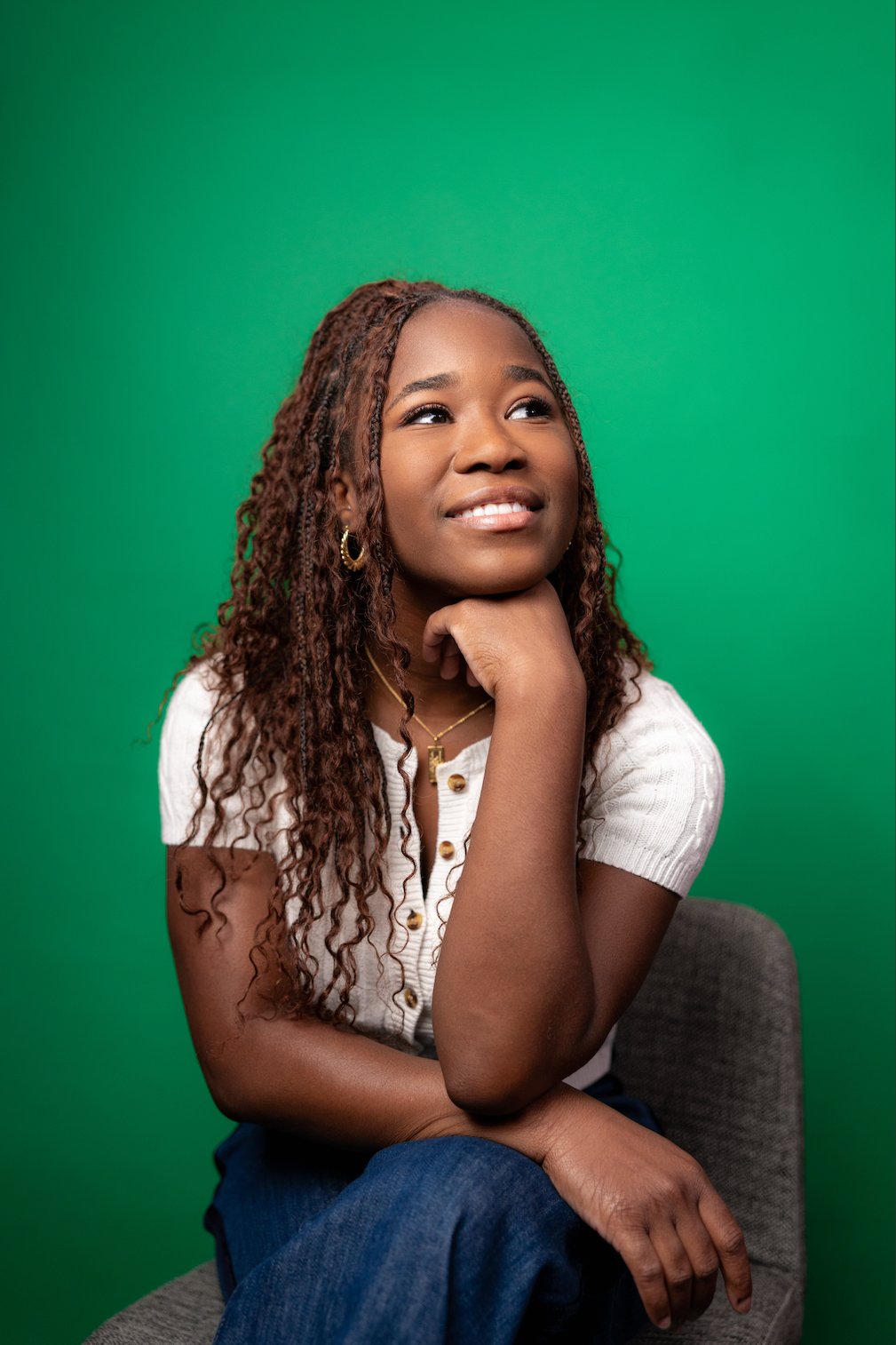 A young woman with curly hair and gold jewelry sitting on a gray chair against a green background, resting her chin on her hand and looking slightly upward.