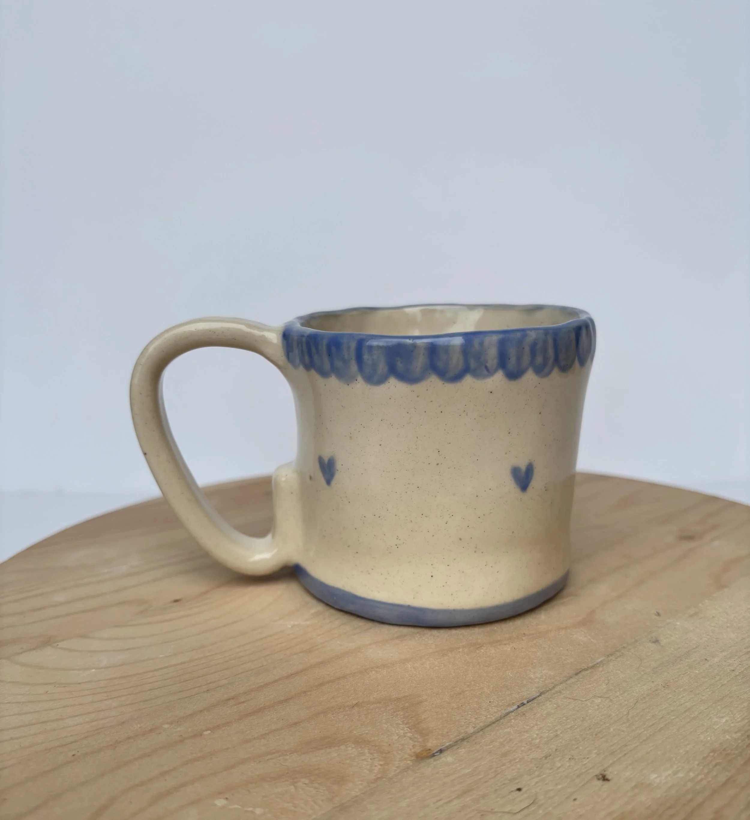 A ceramic mug with a beige background and blue decorative patterns along the rim and small blue hearts on the body, placed on a wooden surface.