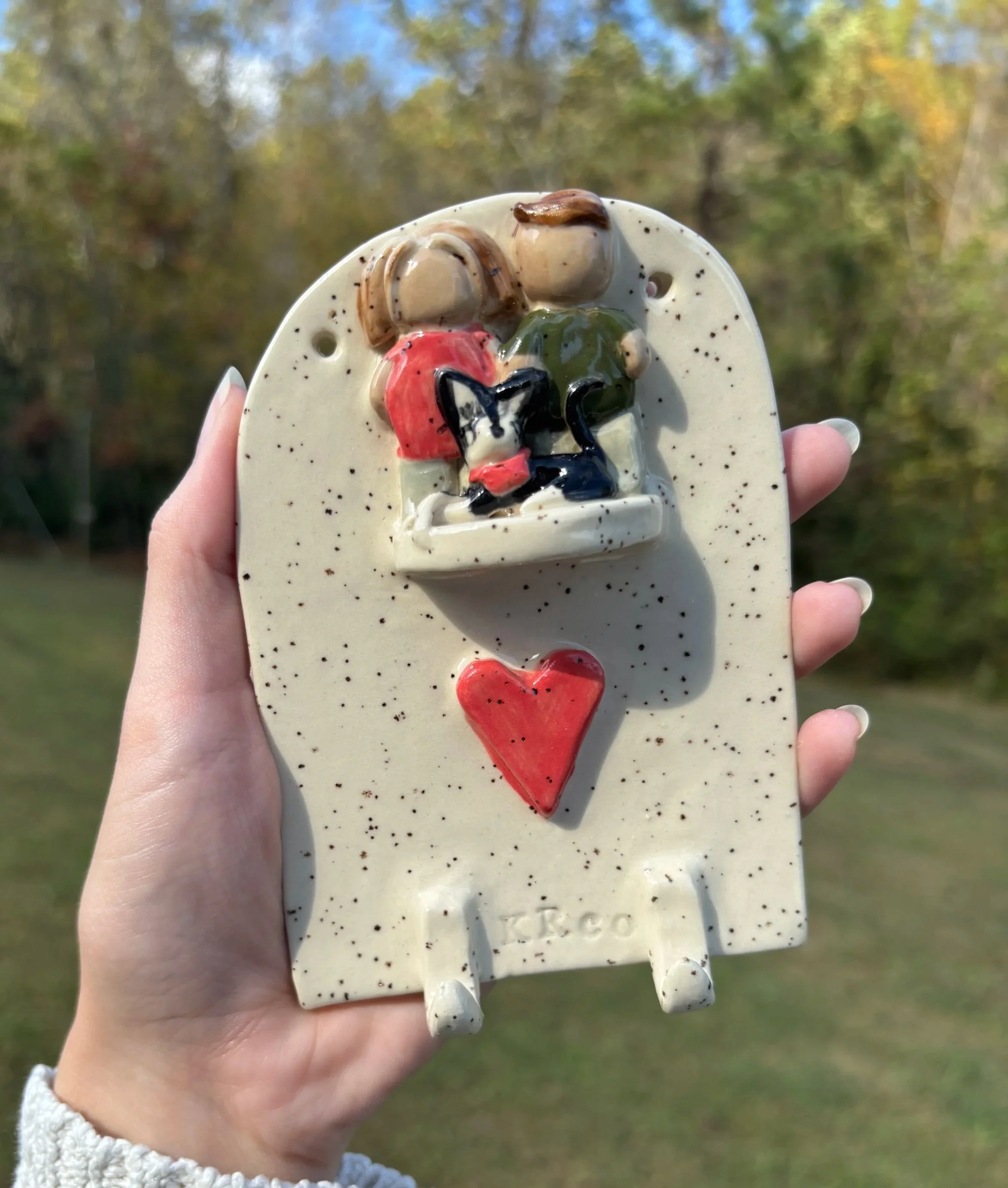 A hand holding a ceramic sculpture of a small group of stylized animals and a red heart on a speckled ceramic base, outdoors with trees in the background.