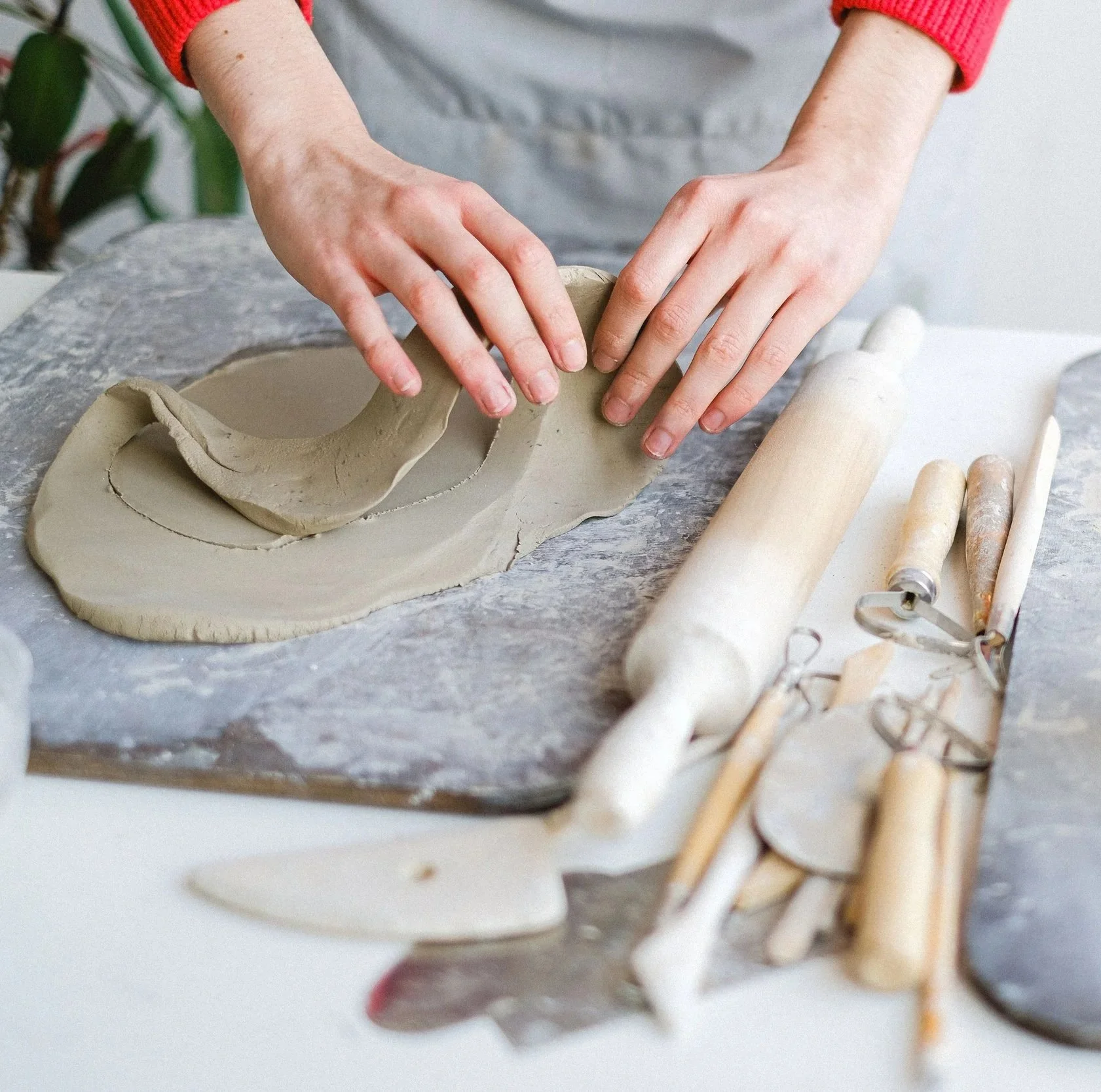 Hands shaping clay on a table, surrounded by pottery tools and a clay sculpture, in a pottery workshop.