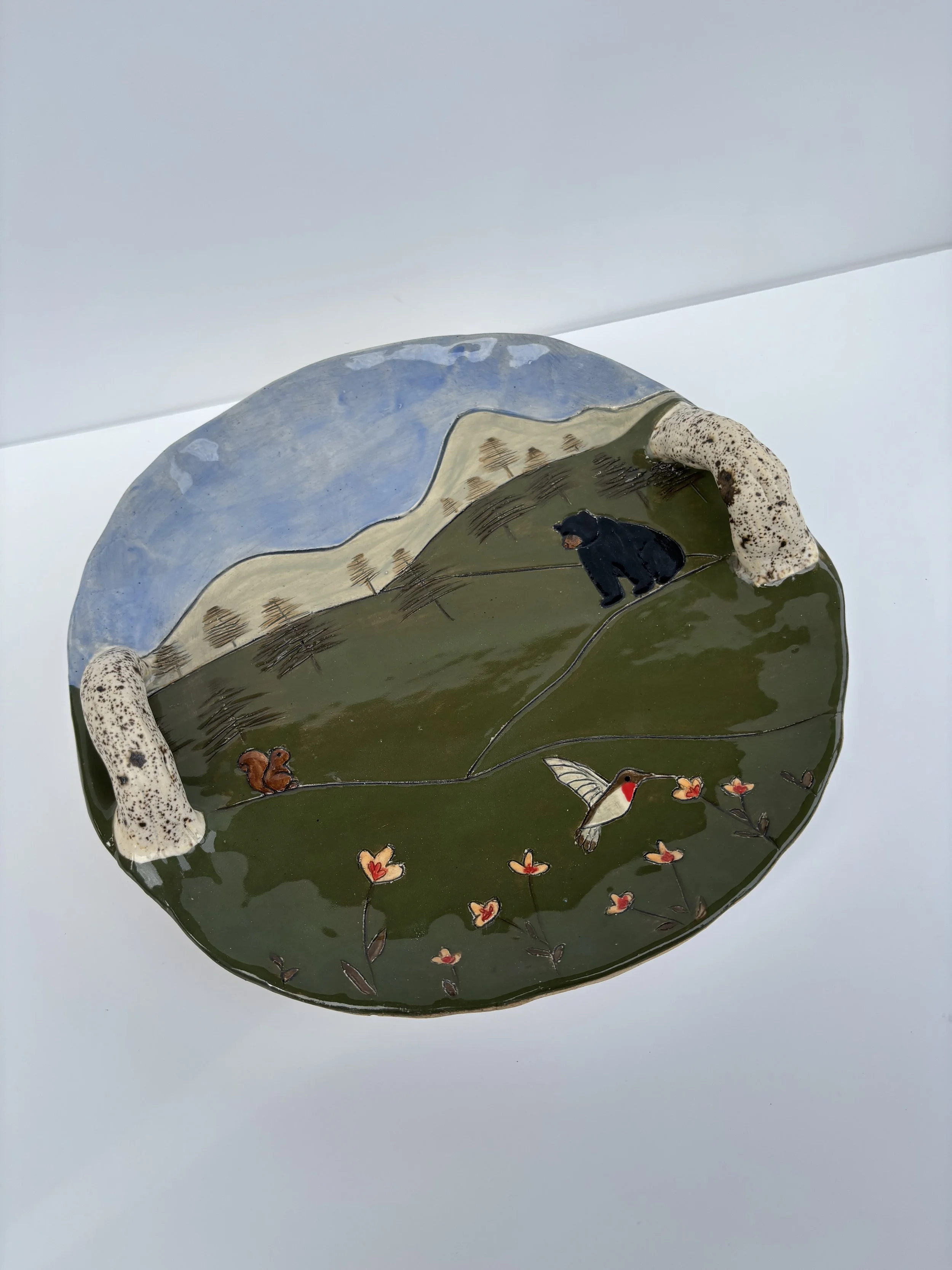 A decorated ceramic plate featuring a landscape scene with mountains, trees, a black bear, a hummingbird, a squirrel, and butterflies, resting on a white surface.