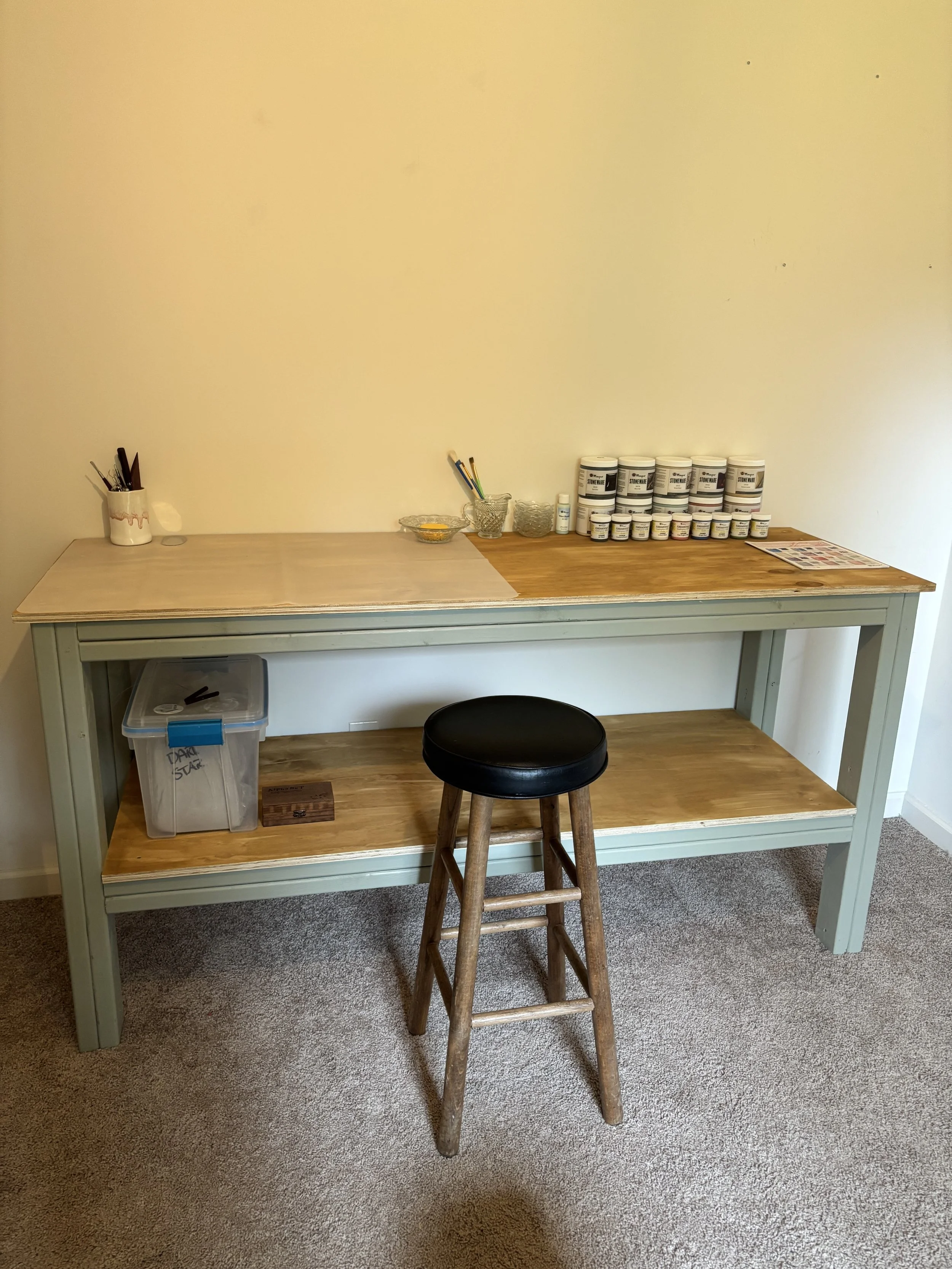 A wooden desk with a pastel green frame and natural wood countertop, holding jars of art supplies, brushes, a small bottle, and a magazine or catalog. There's a wood stool and a plastic storage container underneath, all in a room with beige walls and carpeted floor.