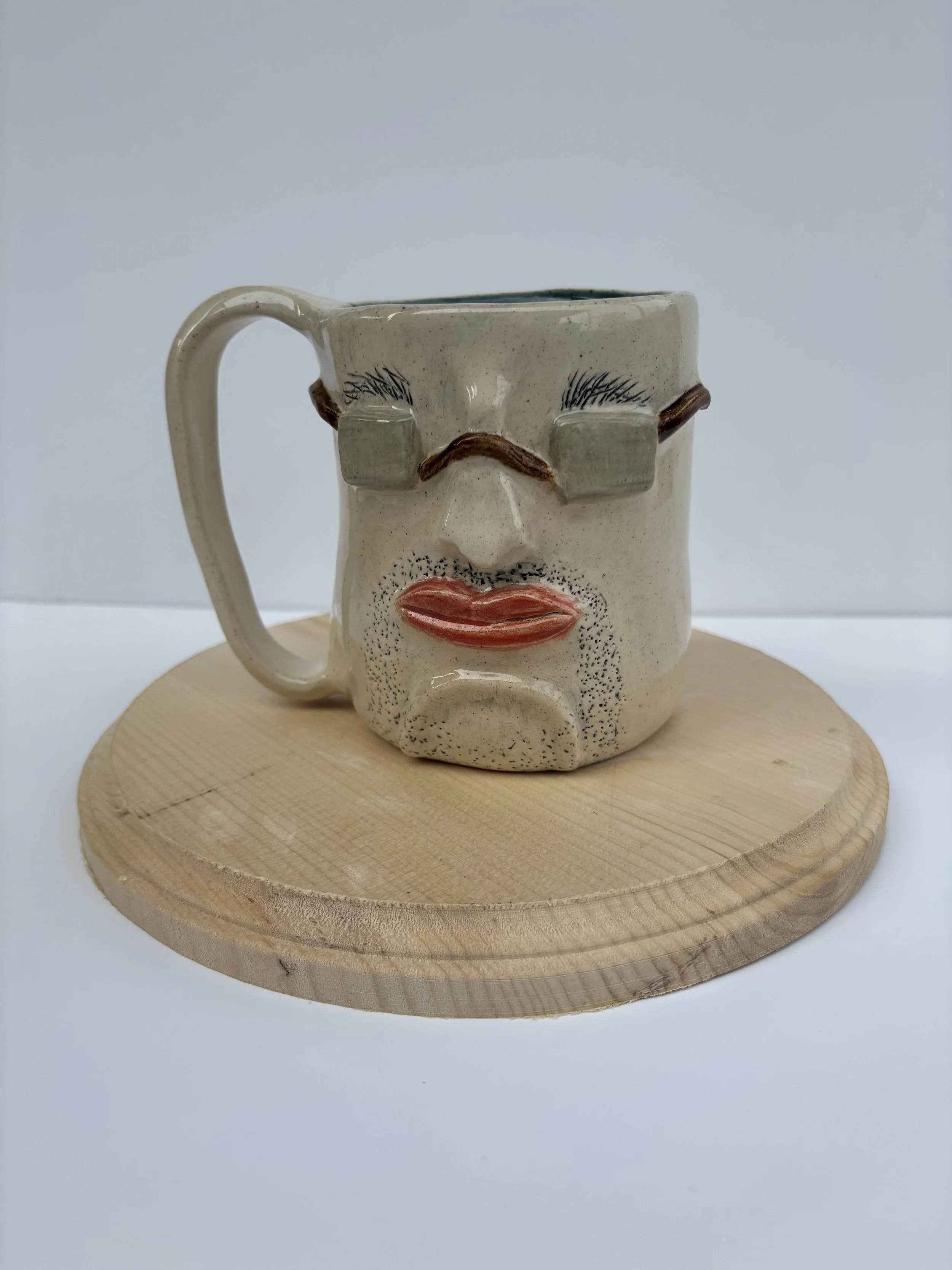 Ceramic mug designed to resemble a human face with exaggerated features, including bushy eyebrows, sunglasses, a nose, and lips, placed on a round wooden stand against a gray background.
