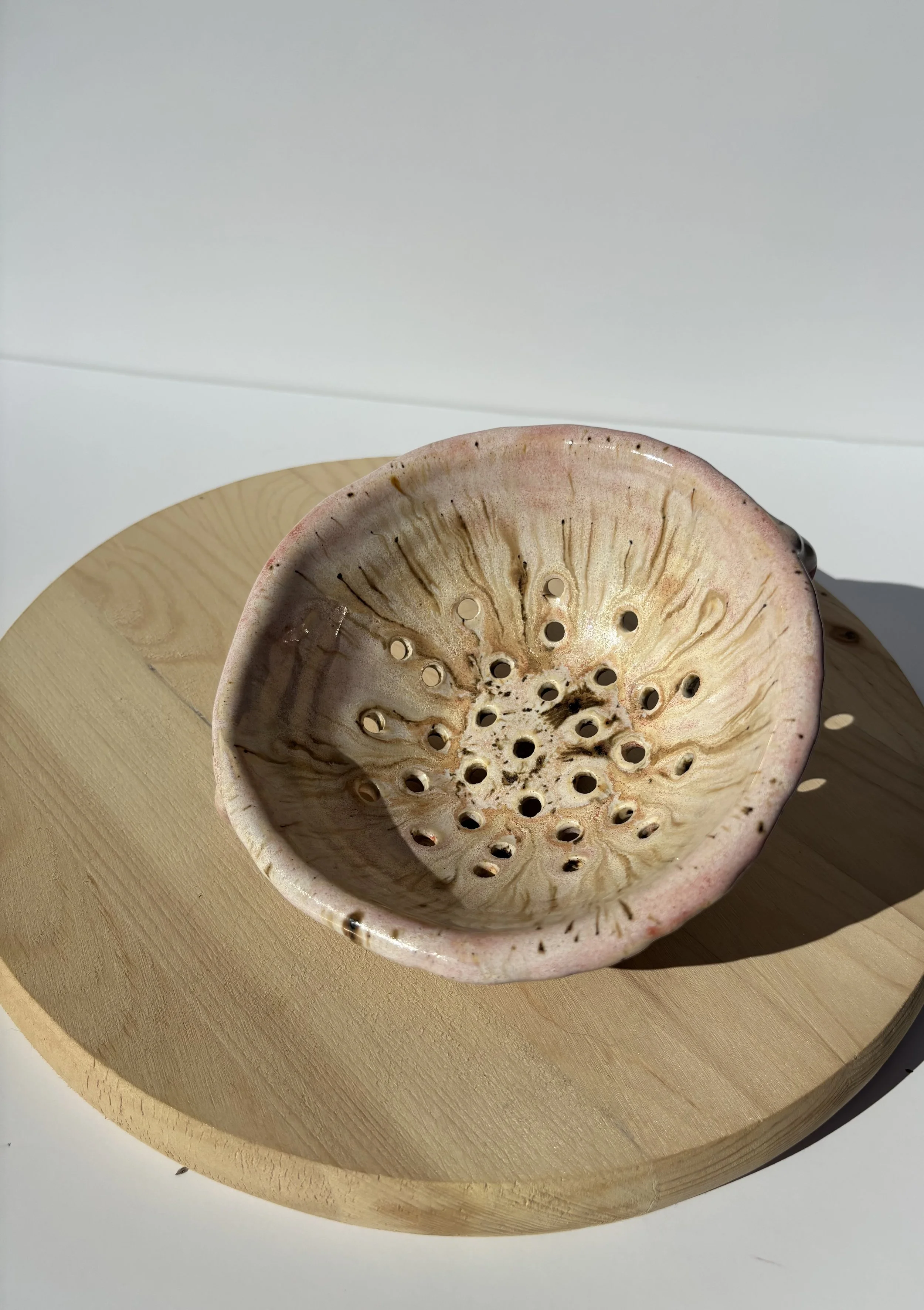 A ceramic colander with holes, placed on a round wooden surface, with a white background.