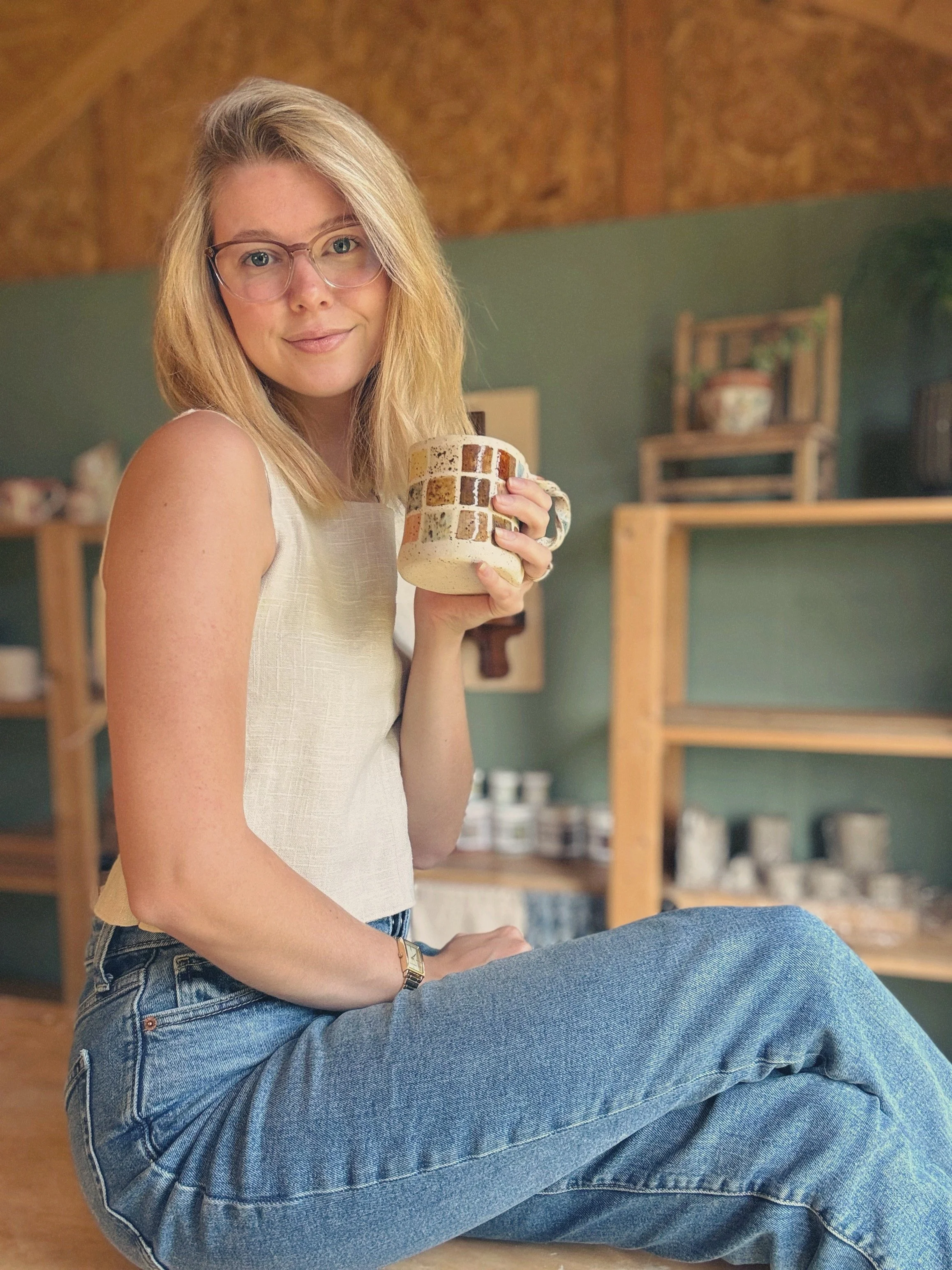 A young woman with blonde hair and glasses smiling, sitting cross-legged and holding a patterned mug, in a cozy room with wooden shelves in the background.