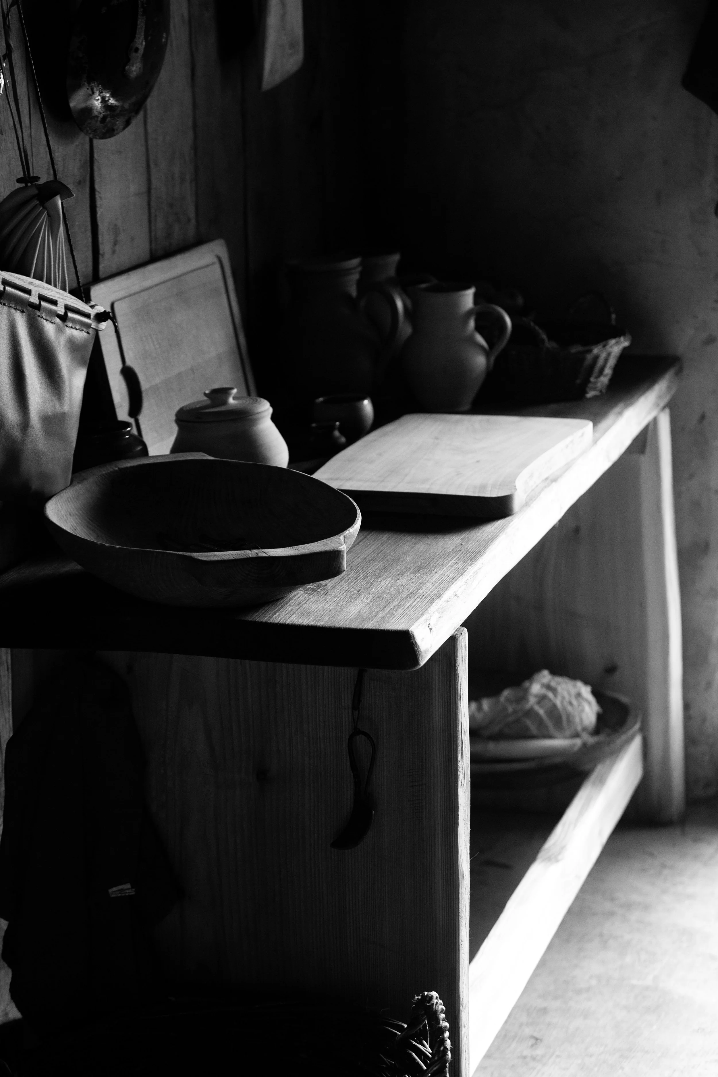 A rustic wooden shelf with various ceramic and pottery items, including a shallow bowl, jars, and vases, against a wood-paneled wall. The scene is in black and white with soft, natural lighting.