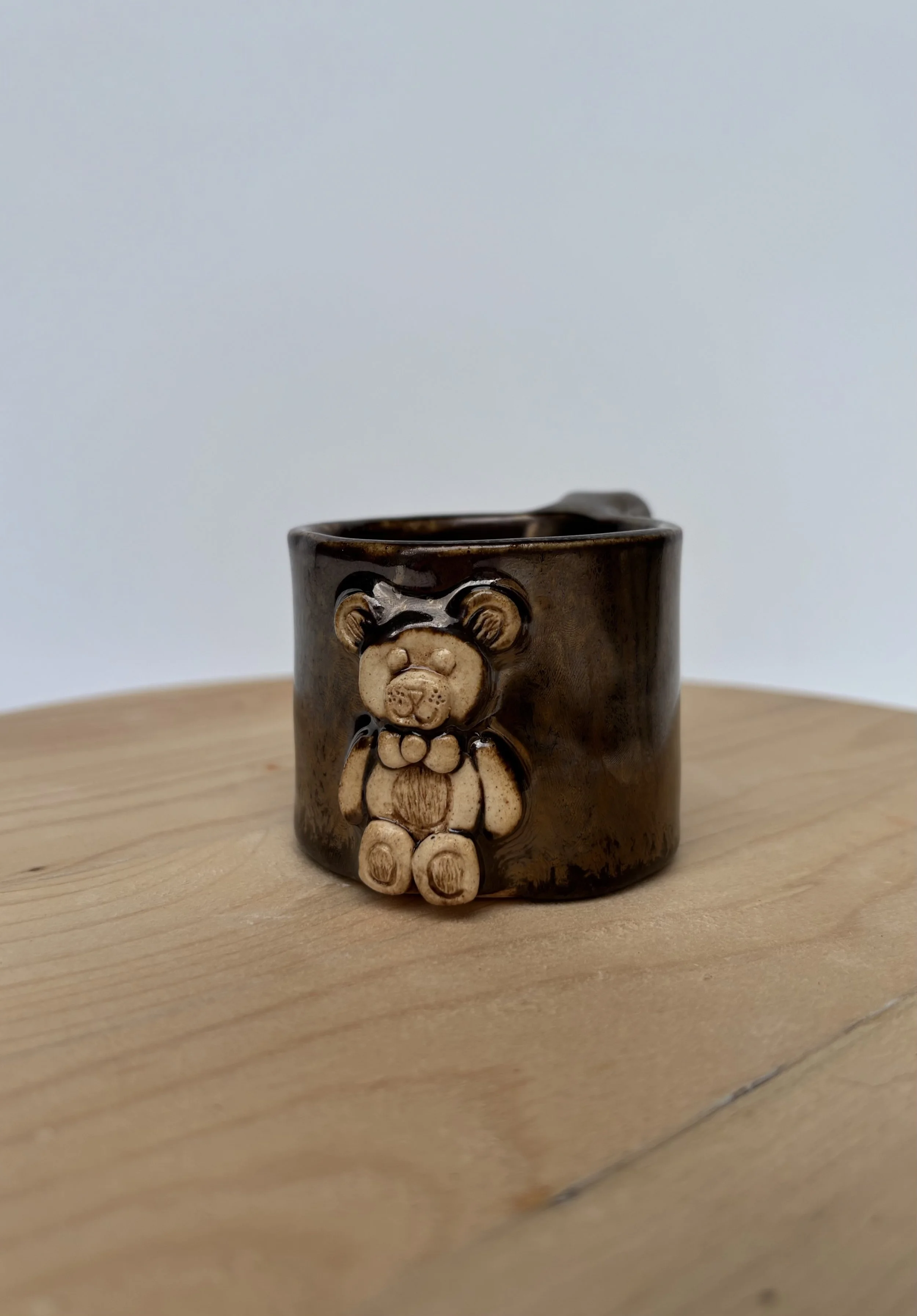 A ceramic mug with a 3D bear figure attached to the front, placed on a wooden surface with a plain light-colored background.