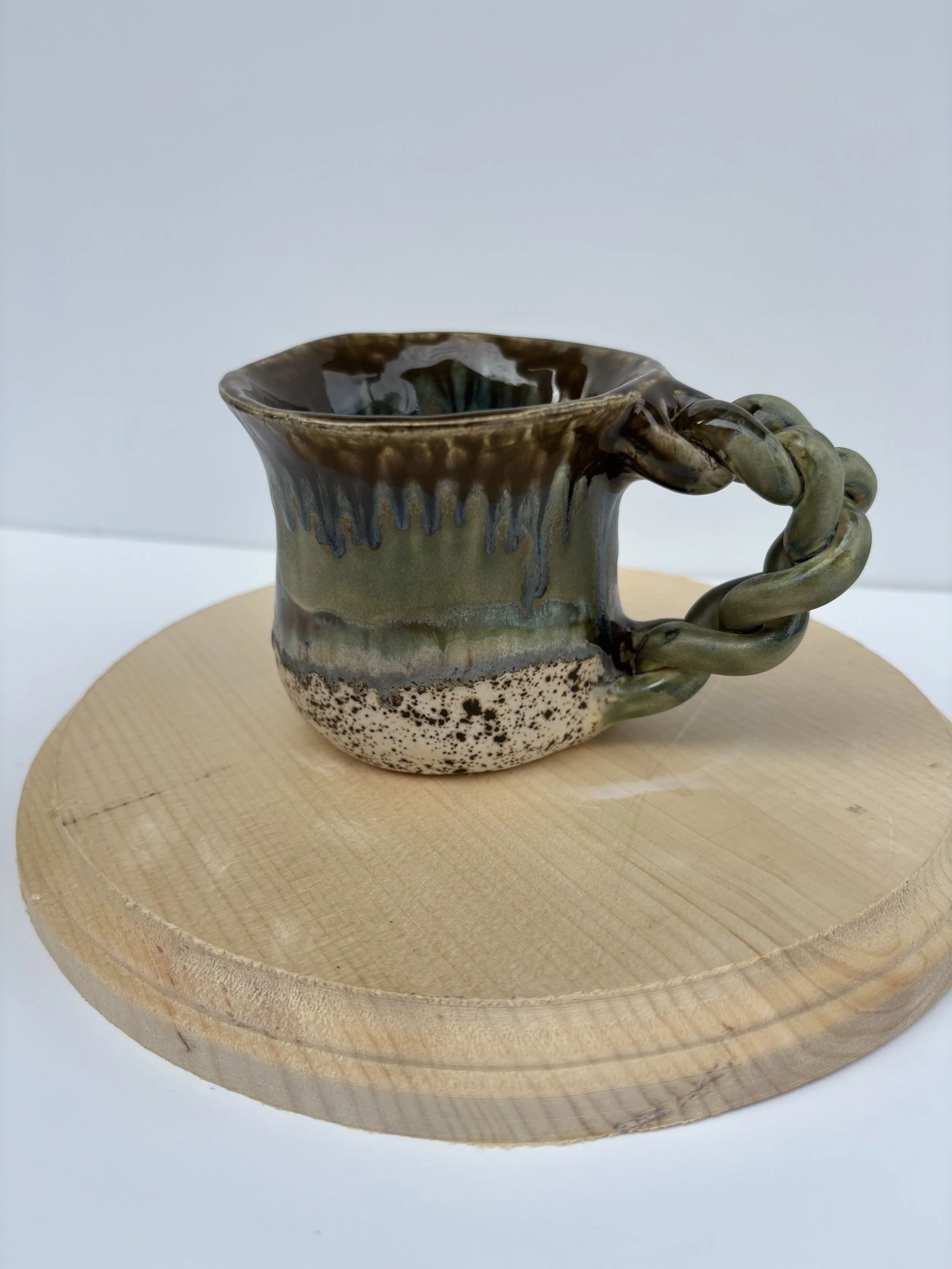 Ceramic mug with a glazed, brown and green design on a wooden circular platform.