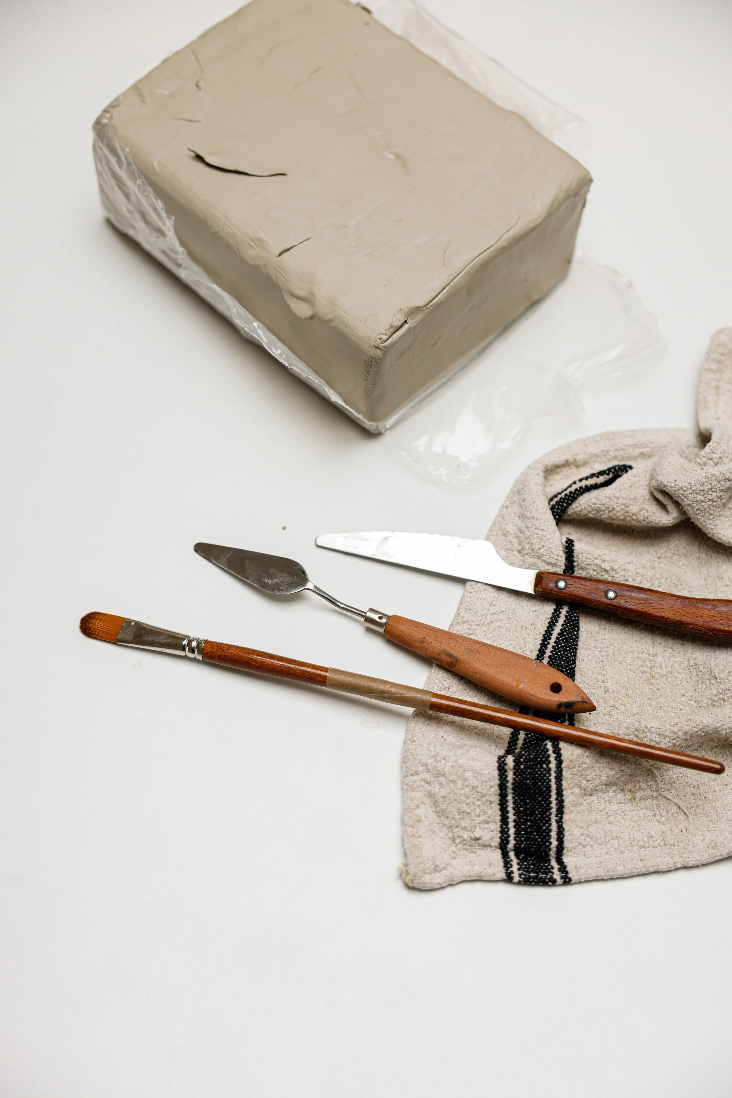 A block of clay wrapped in plastic, lying on a white surface. Next to it are two sculpting tools with wooden handles, one with a flat, wide blade and the other with a pointed, curved blade. There is also a towel with black stripes.
