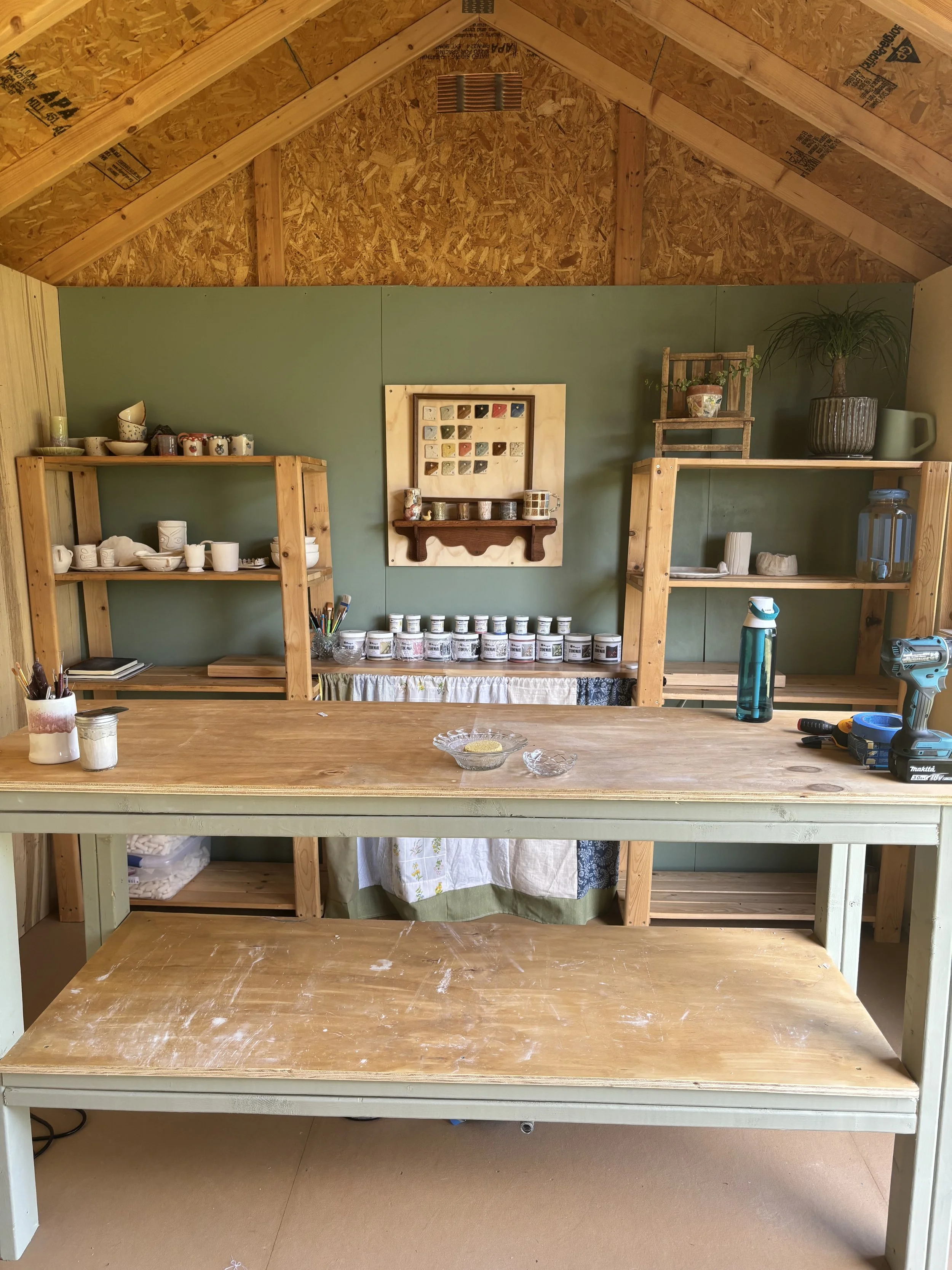 Inside a workshop with unfinished wooden shelving, bottles and tools on the shelves, a greenish table with some items on it, and a partially painted wall with a decorative framed piece.