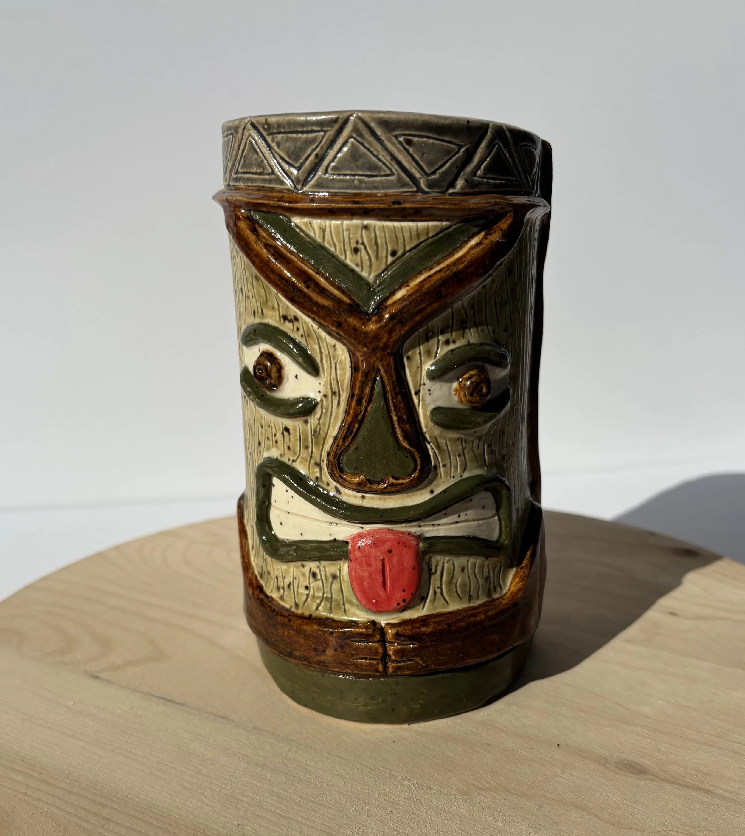 Colorful ceramic tiki trophy with a smiling face, red tongue, and geometric patterns on the top, sitting on a wooden surface against a plain background.