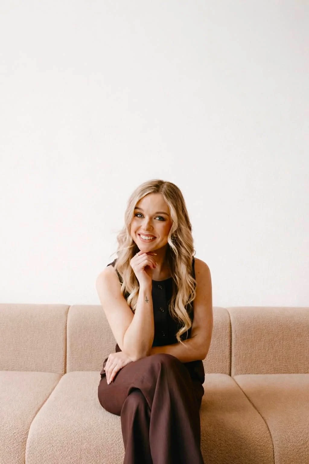 A young woman with long blonde wavy hair sitting on a beige sofa, smiling, wearing a black sleeveless top and brown pants, with a white plain wall in the background.