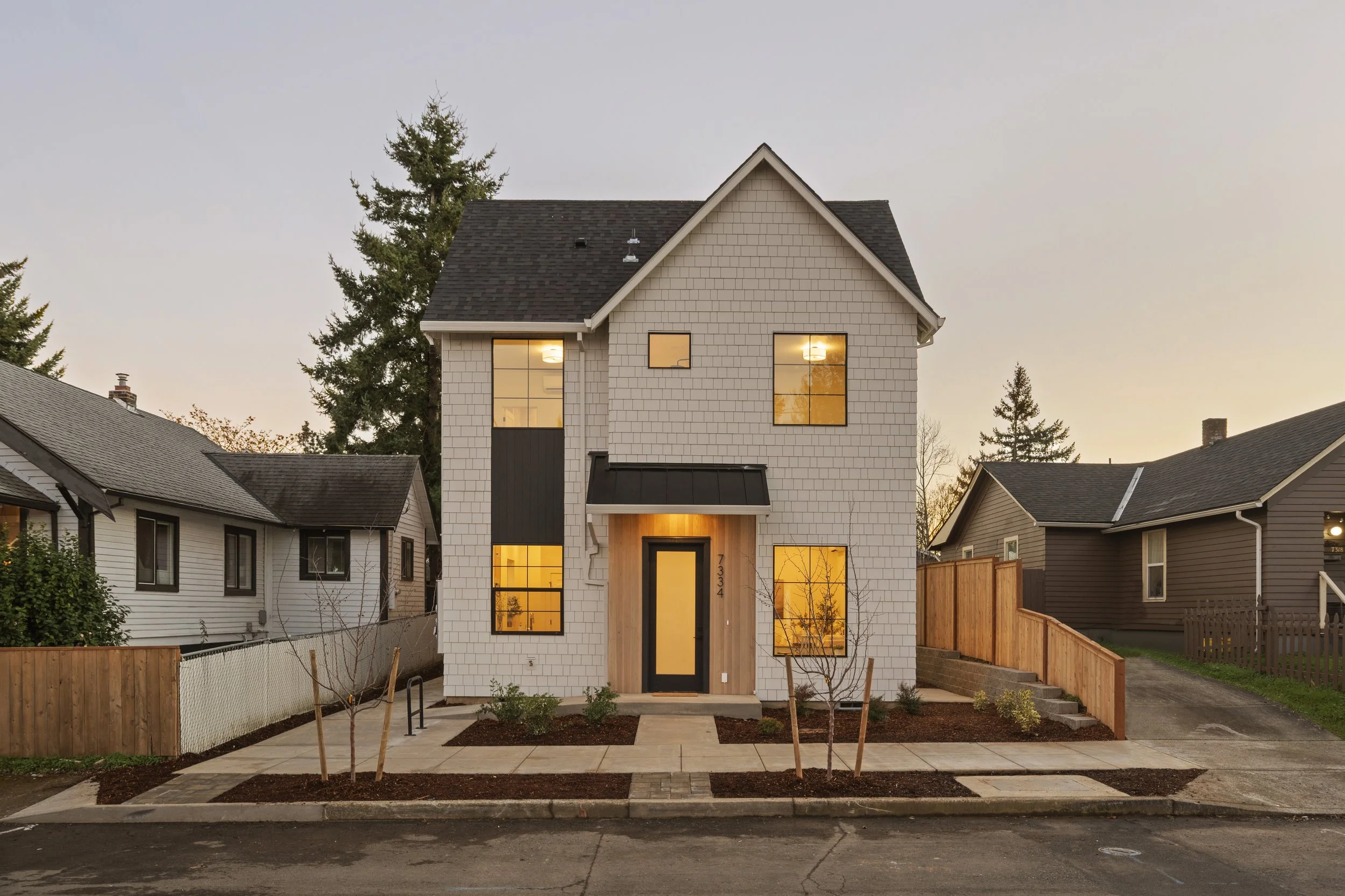 A modern white house with black trim and large windows, situated in a neighborhood with similar homes. The house has a small front yard with newly planted trees and a paved walkway, surrounded by a wooden fence and neighboring houses.