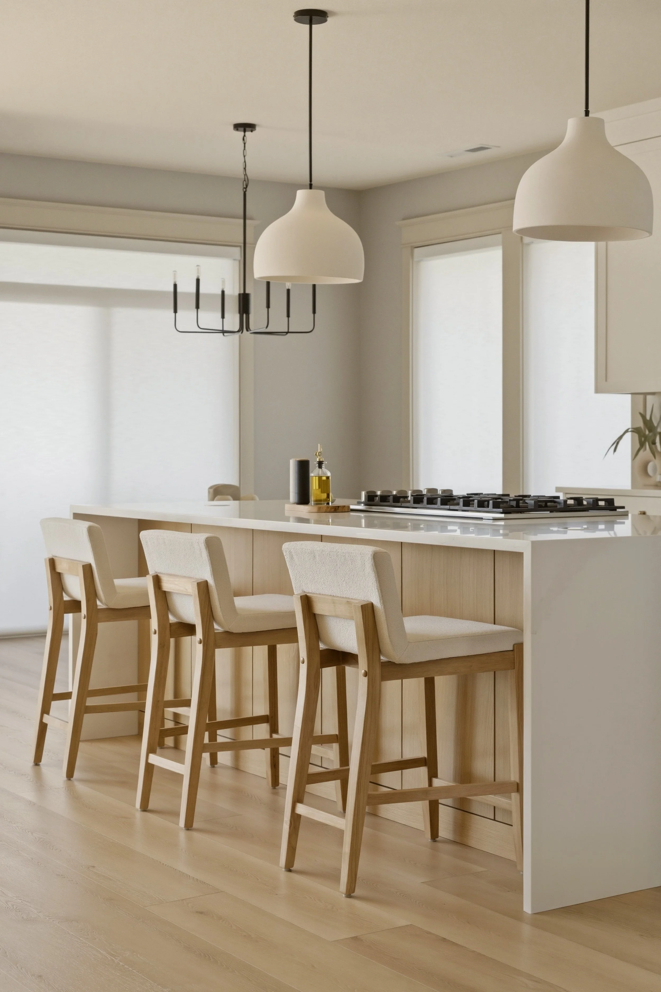 A modern kitchen with a white island counter, four wooden bar stools with light upholstered seats, white pendant lights, and windows with white blinds, all in a bright and minimalist style.
