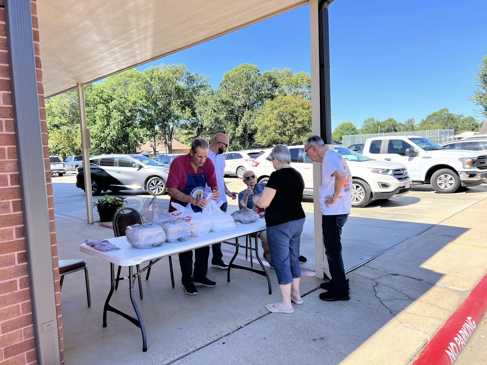 A group of people gathered around a table outside of a building, possibly at a community event, with several parked cars and green trees in the background on a sunny day.