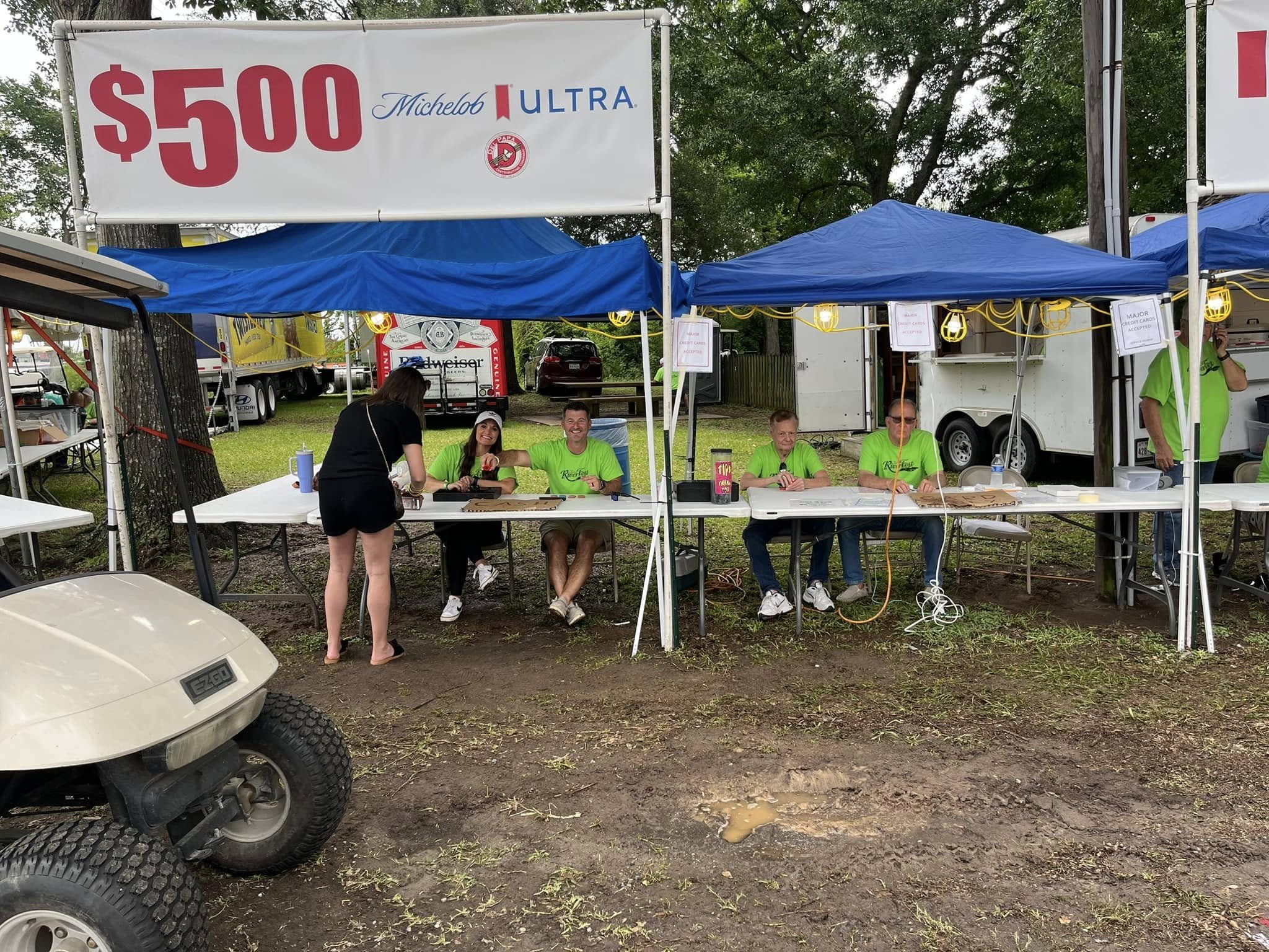 A group of people sitting and standing under blue canopies at an outdoor event, with a large sign advertising $500 Michelob Ultra, and trees in the background.
