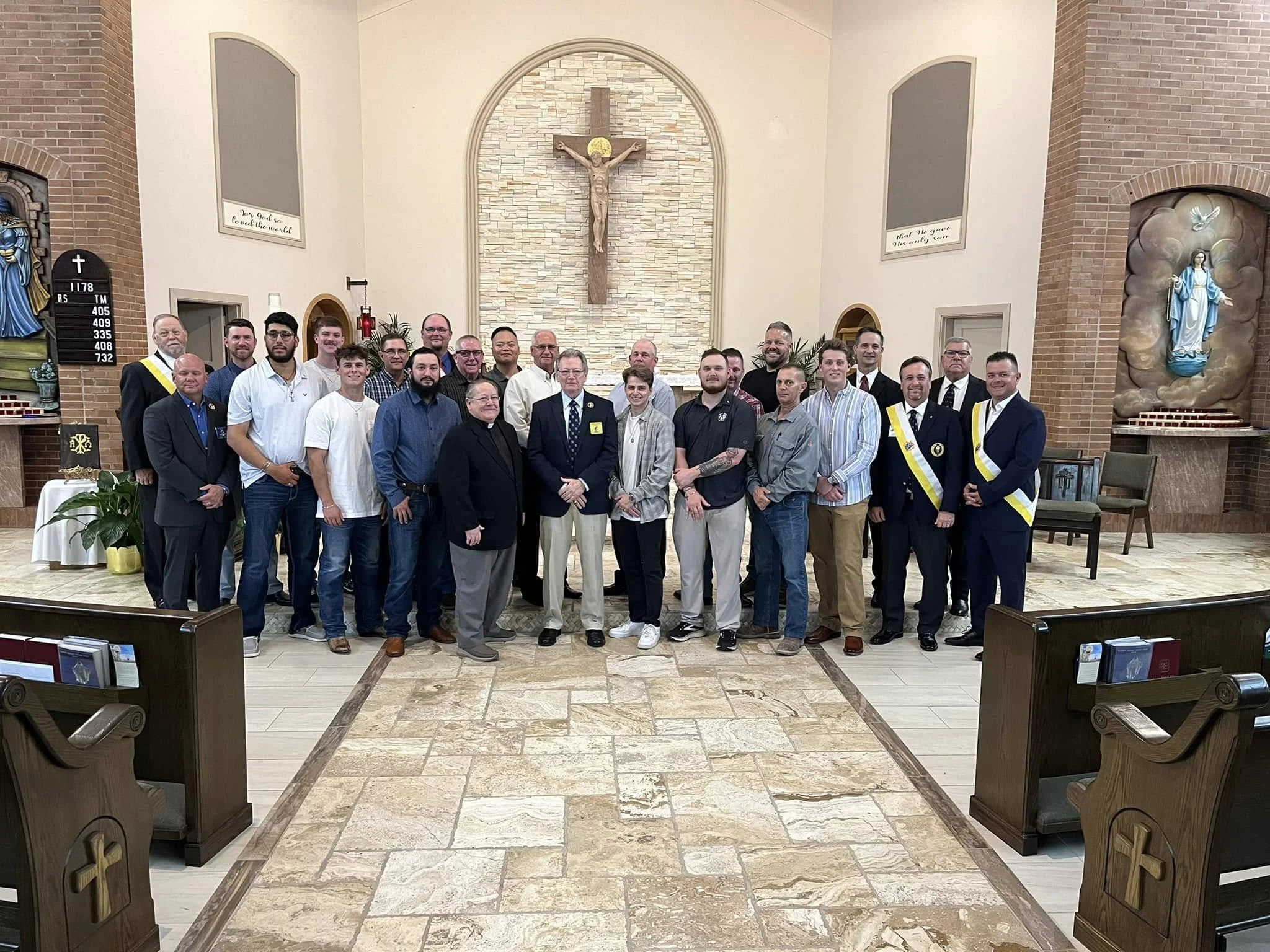 A group of people standing inside a church near the altar, with a large crucifix on the wall behind them, flanked by religious statues and artwork on the walls.