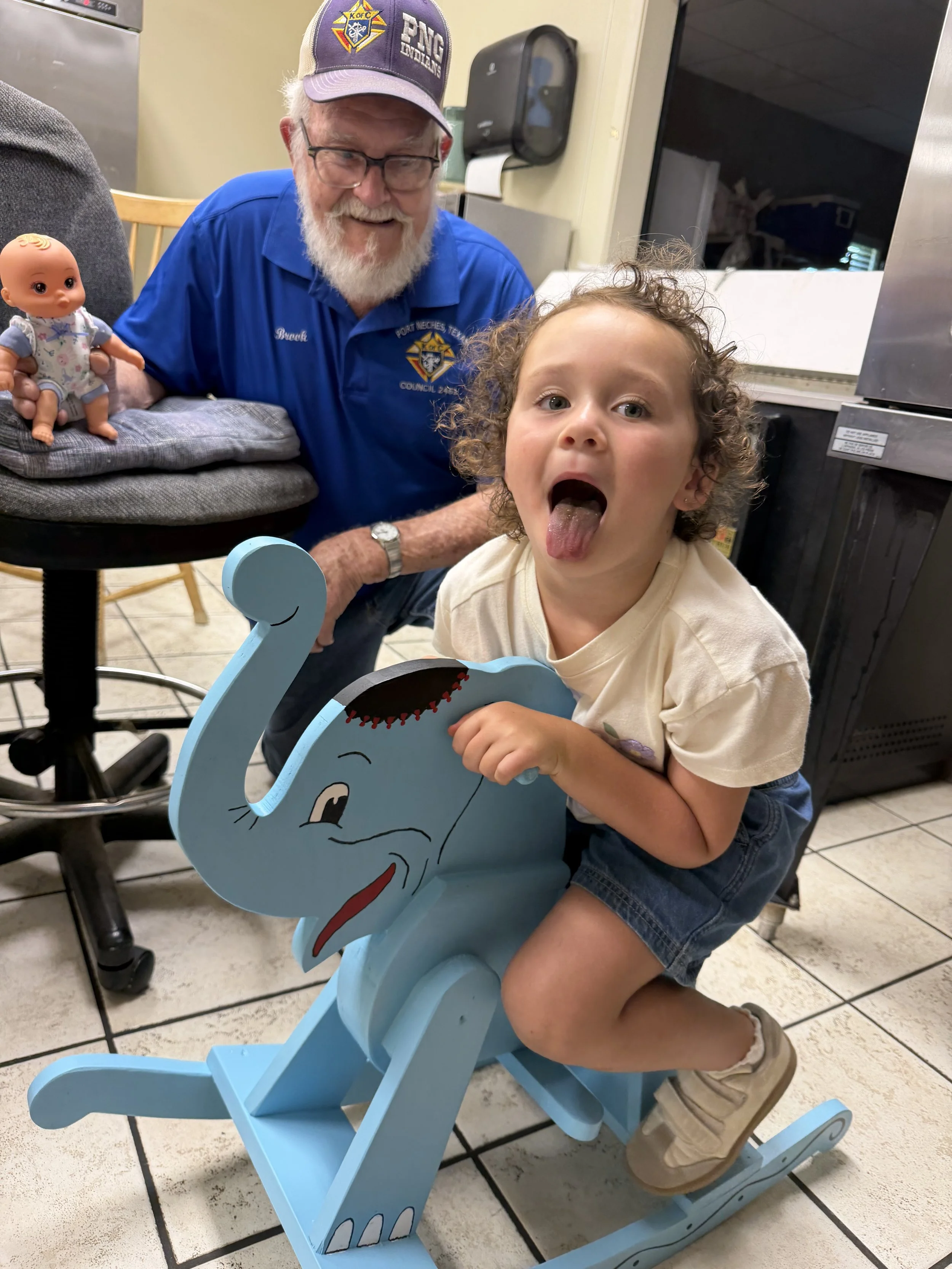 An elderly man with white hair, glasses, and a beard wearing a blue shirt and a purple hat, is smiling and looking at a young girl with curly hair showing her tongue. The girl is sitting on a blue wooden rocking elephant, and there is a doll on a cha