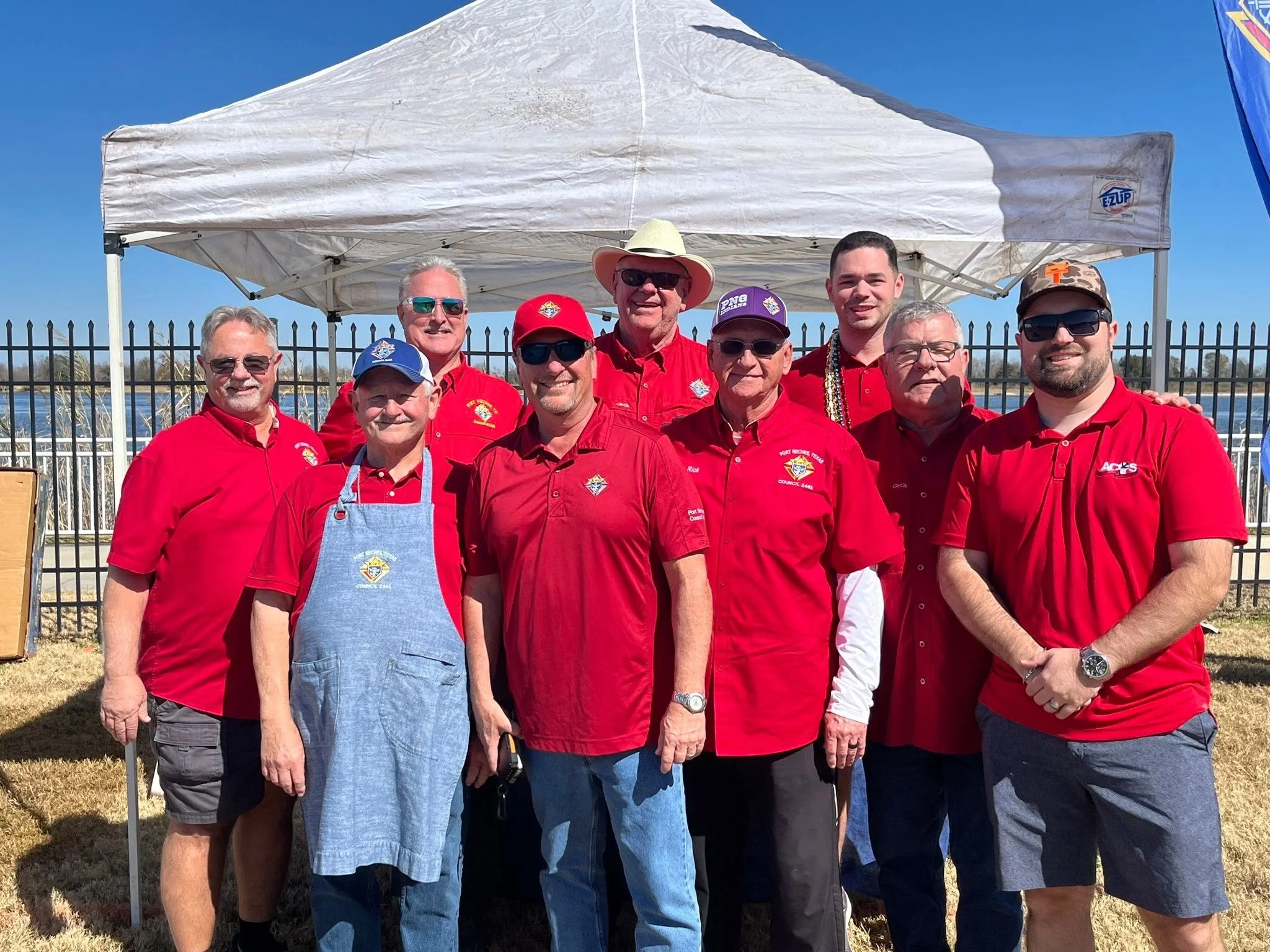 Group of ten men in red shirts and hats standing outdoors in front of a white canopy tent, with a lake and blue sky in the background.