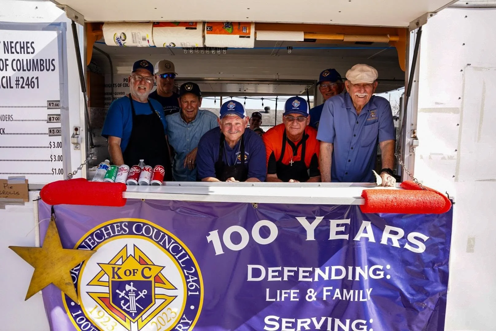 Group of older men working at a vendor booth with a purple banner celebrating 100 years, affiliated with Knights of Columbus, selling bottled drinks and food at an event.