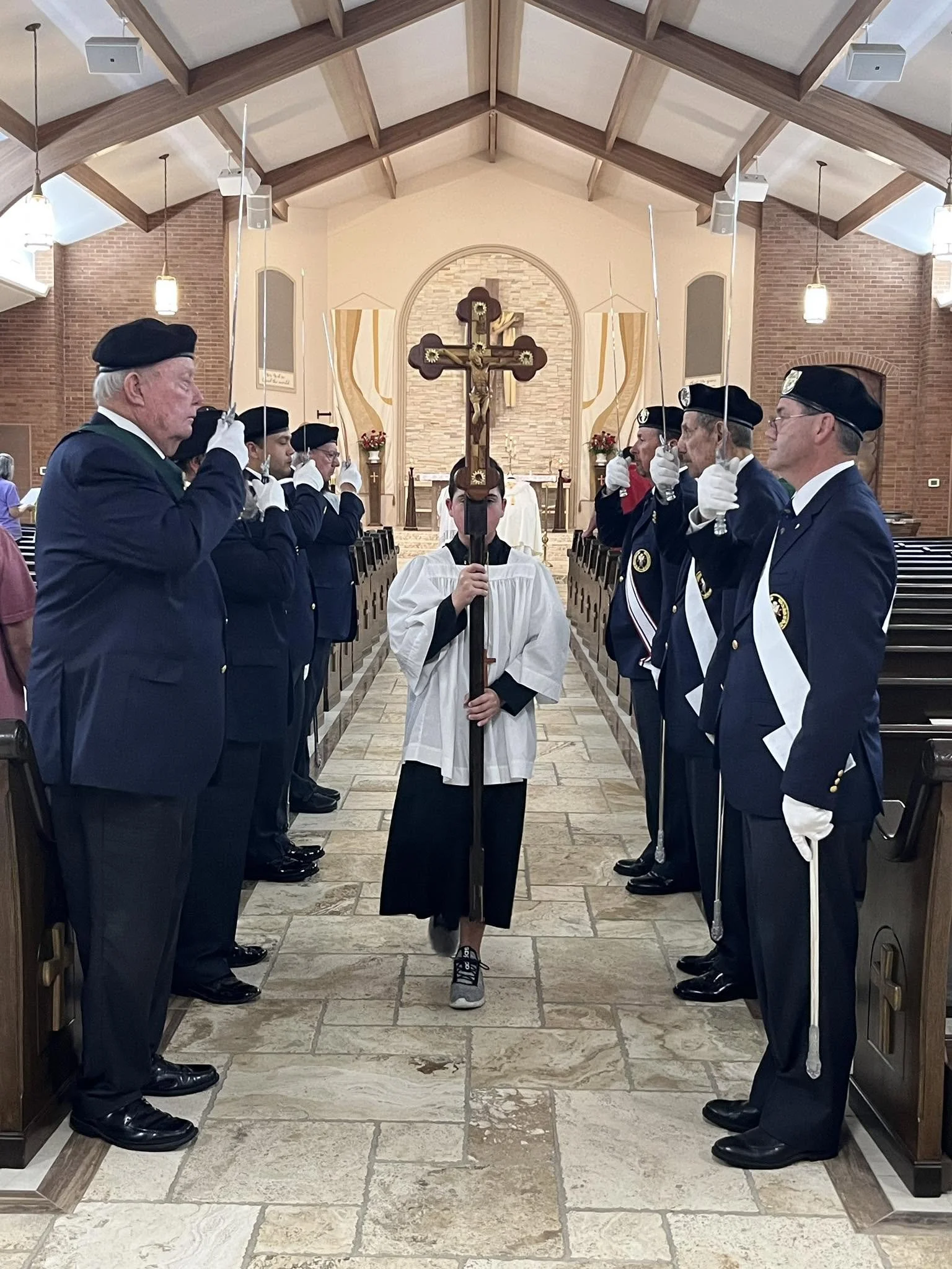 A church interior with a person holding a cross and standing in front of an altar, surrounded by people in uniform holding swords. The setting has a vaulted wooden ceiling, brick walls, and chandeliers.