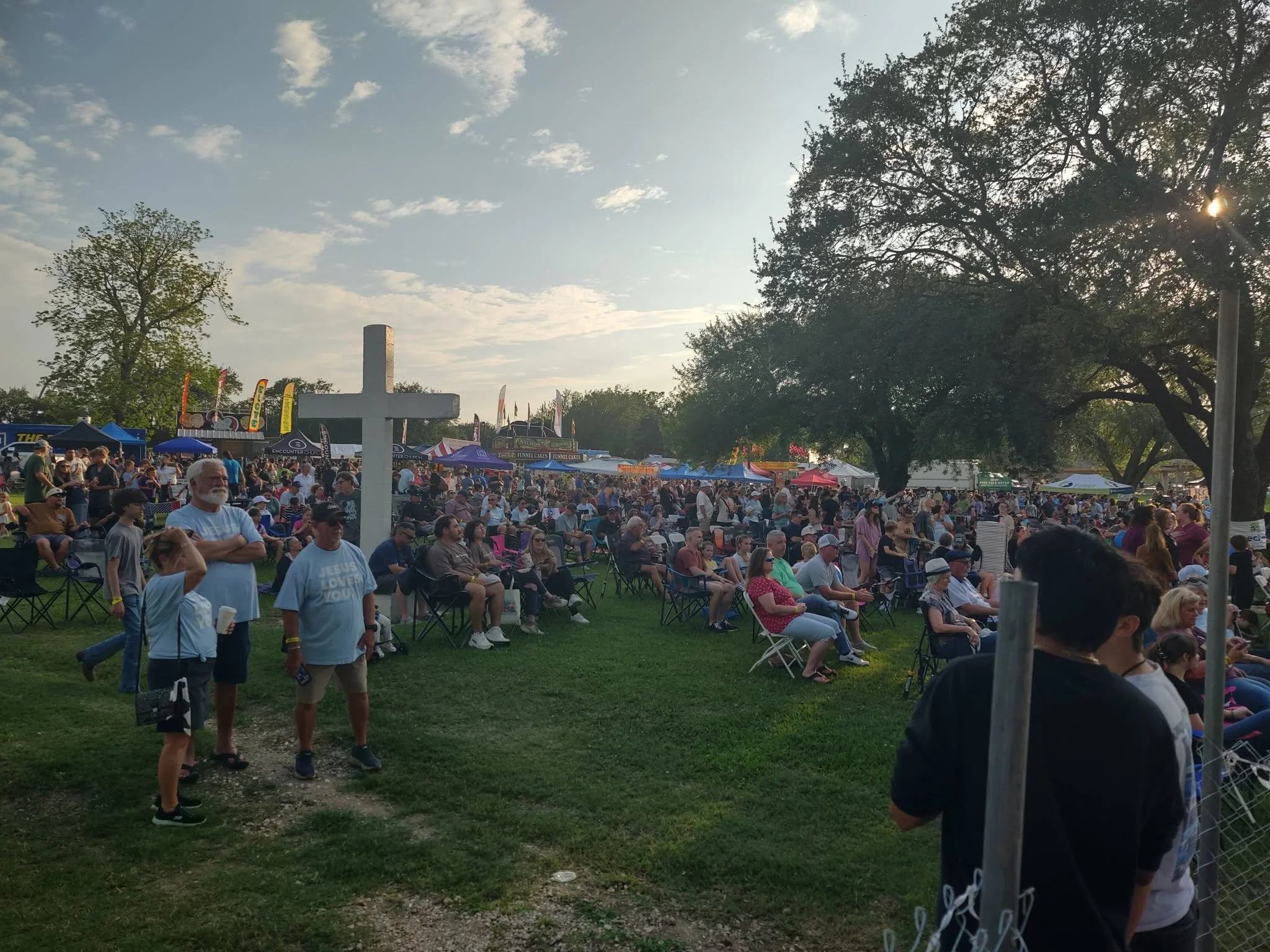 A large crowd of people gathered outdoors at a festival or fair, with tents and food stands visible. The scene is set during late afternoon or early evening, with trees and blue sky in the background.