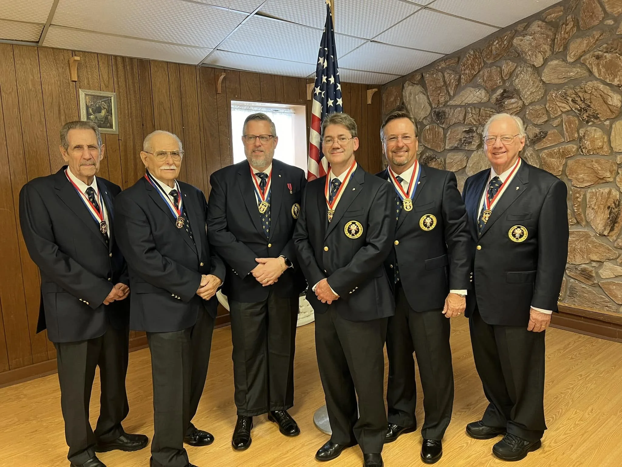 A group of six older men in suits with medals around their necks standing in a room with wooden panel walls and a stone accent wall, with an American flag and a small painting behind them.