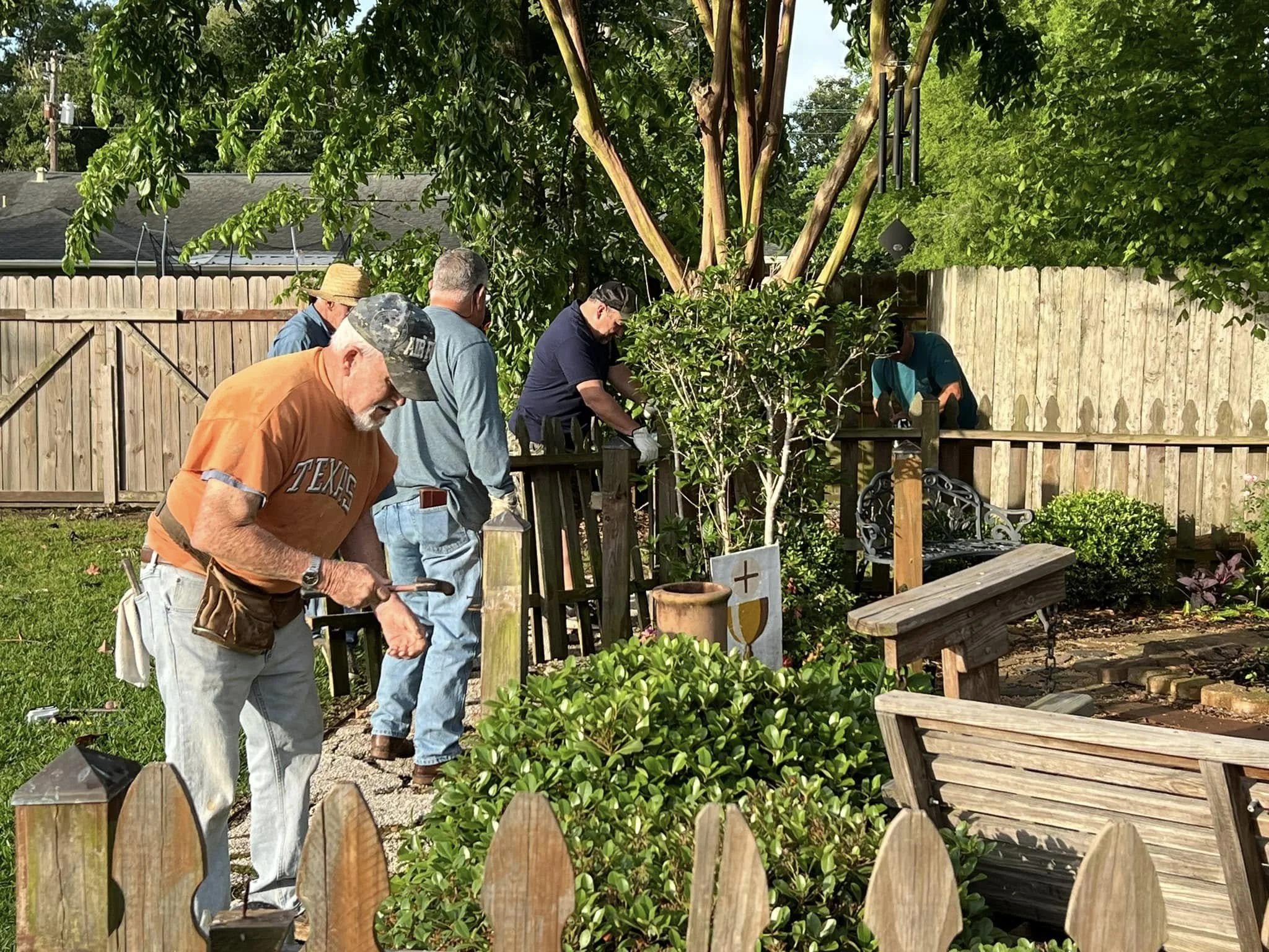 A group of people working together in a backyard garden or outdoor area, surrounded by greenery, with a wooden fence in the background and various gardening tools and decorations nearby.