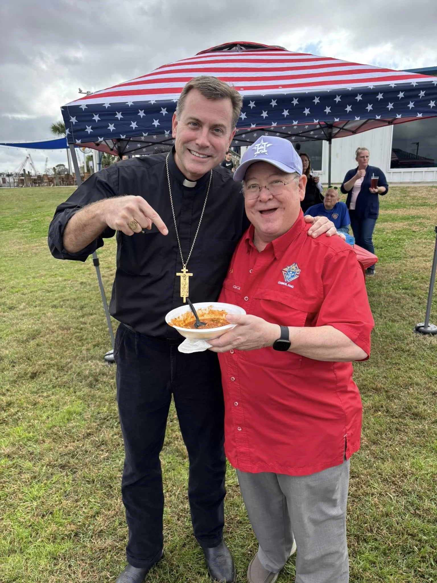 Two men, one dressed as a priest and the other in a red shirt, smiling and posing under a striped American flag tent at an outdoor gathering. The priest is pointing at the man in red, who is holding a bowl of food.