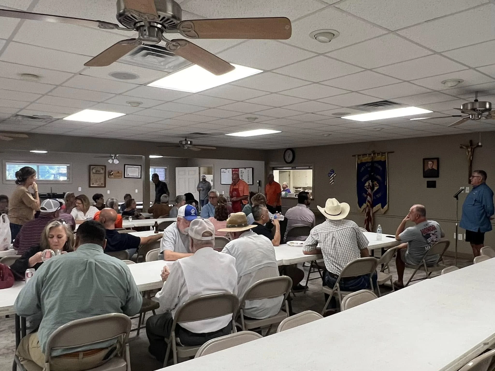 A large room filled with people seated at tables attending a meeting or event. Several individuals are standing at the back, and some are speaking at a podium. The room has a white ceiling with grid pattern, ceiling fans, and fluorescent lighting. Th
