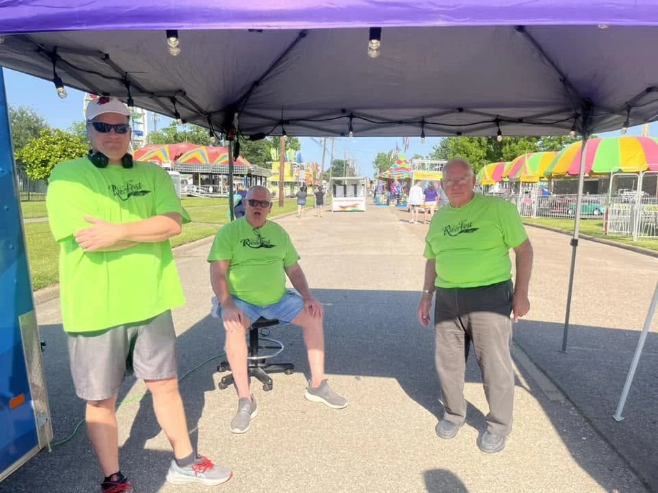 Three men in bright green shirts standing under a tent at a fairground with carnival rides and colorful umbrellas in the background.