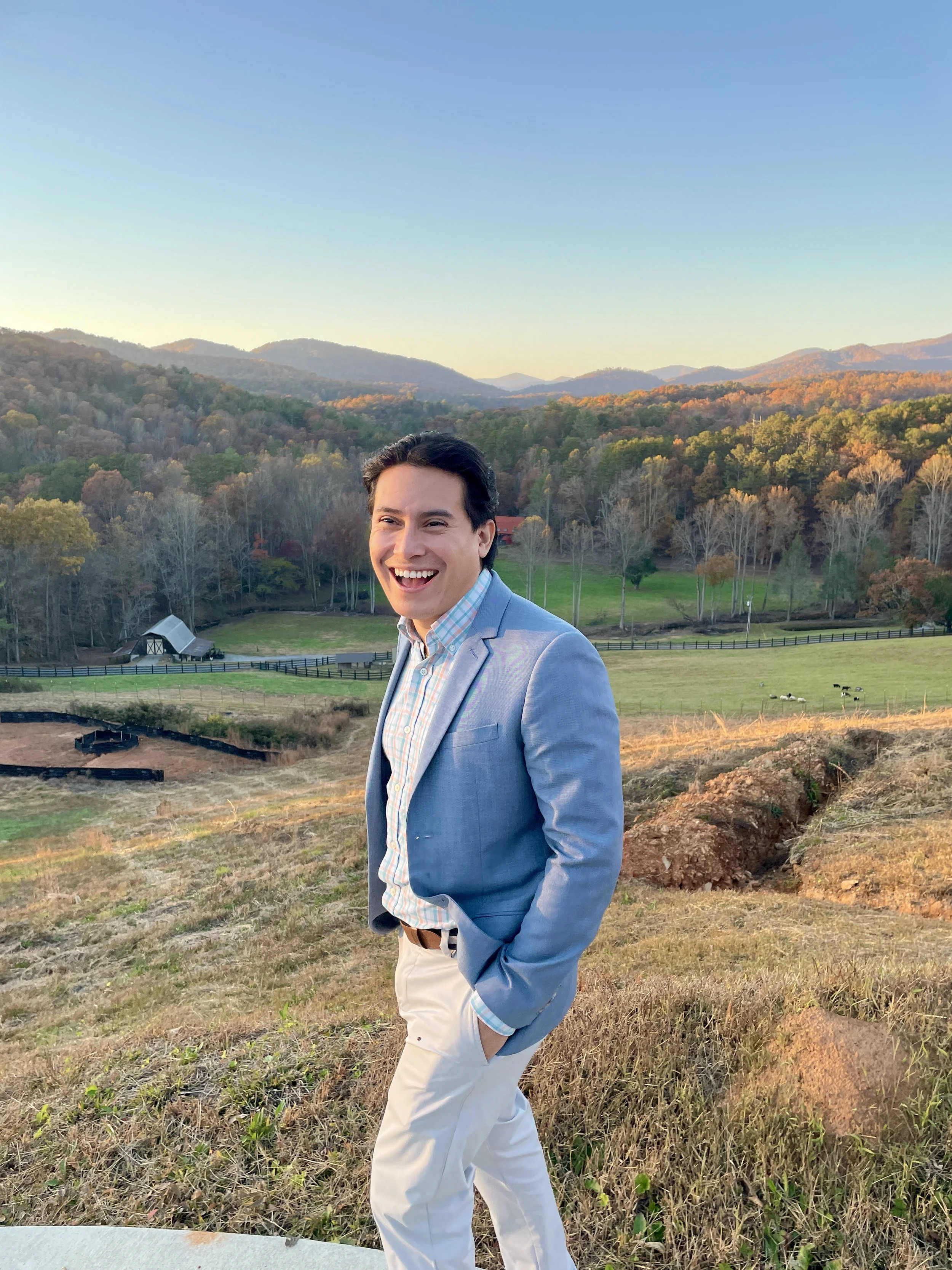 A smiling man in a light blue blazer and white pants standing outdoors on a grassy hillside with a farm and wooded hills in the background during sunset.