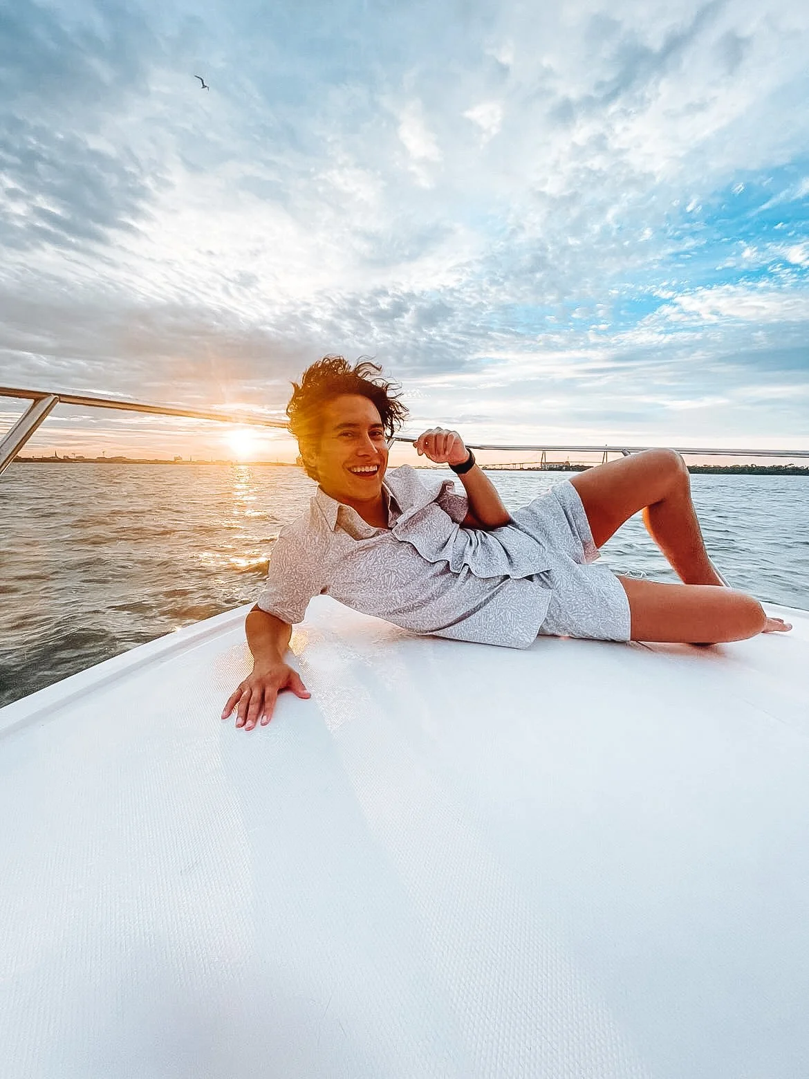 A young man relaxing on the bow of a boat during sunset, smiling at the camera with the water and sky in the background.