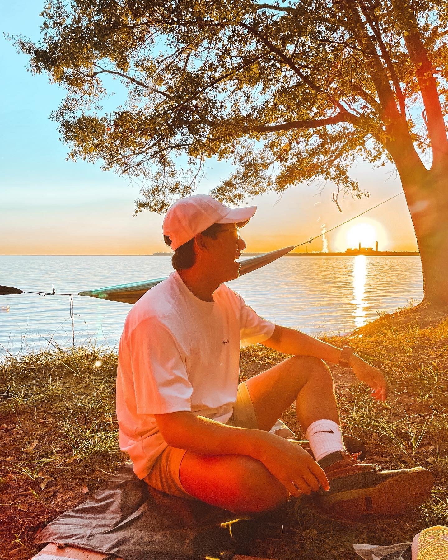 A young man in a white t-shirt, orange shorts, and white Nike socks, sitting on the ground by a lake at sunset, holding a fishing rod with a fish caught on the line. There is a large tree overhead and a kayak in the background, with factory smokestacks visible on the horizon.