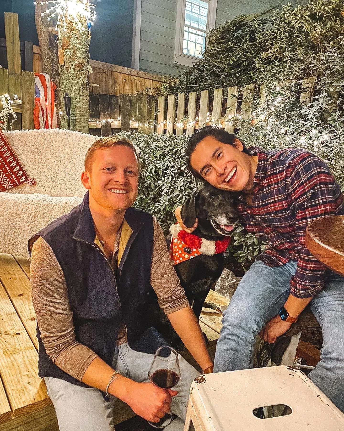 Two men and a black dog with a red sweater sitting outdoors at night. One man has short blonde hair and is smiling, holding a glass of red wine. The other man has dark hair tied back, is wearing a plaid shirt, and is leaning towards the dog. The setting features a wooden deck, string lights, and bushes, creating a cozy outdoor atmosphere.