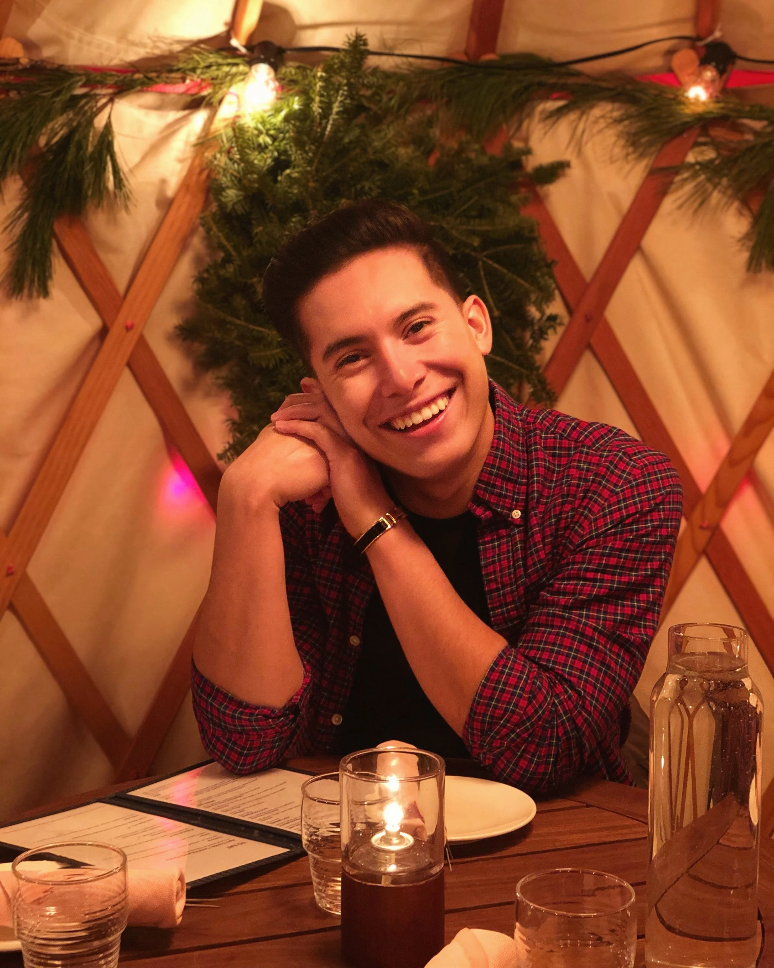 A smiling young man with dark hair, wearing a red and black plaid shirt, sitting at a wooden table with glasses of water and a lit candle. The background is decorated with pine branches and lights, creating a festive or cozy atmosphere.