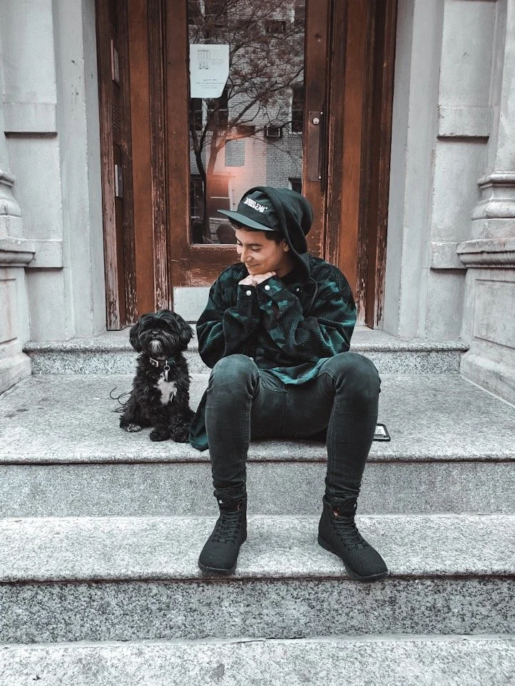 A young person with short hair, wearing a black hat, a dark plaid shirt, and black pants, is sitting on stone steps outside a building with a wooden door. They are smiling and looking at a small black dog sitting next to them.