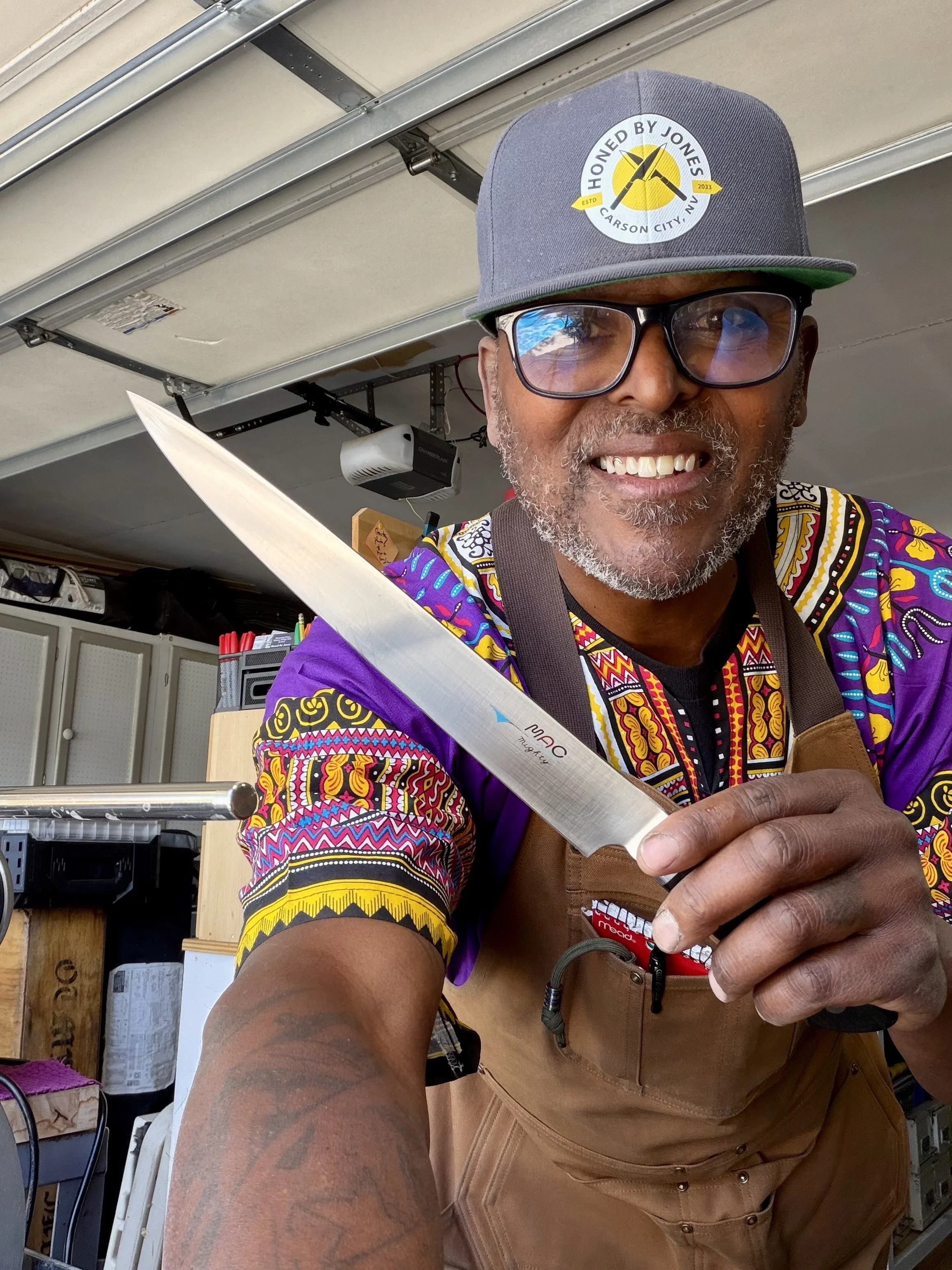 A man smiling and holding a kitchen knife, wearing glasses, a gray cap with a logo, and a colorful shirt with black, yellow, red, blue, and white patterns. He is in a workshop or garage with shelves, tools, and equipment in the background.