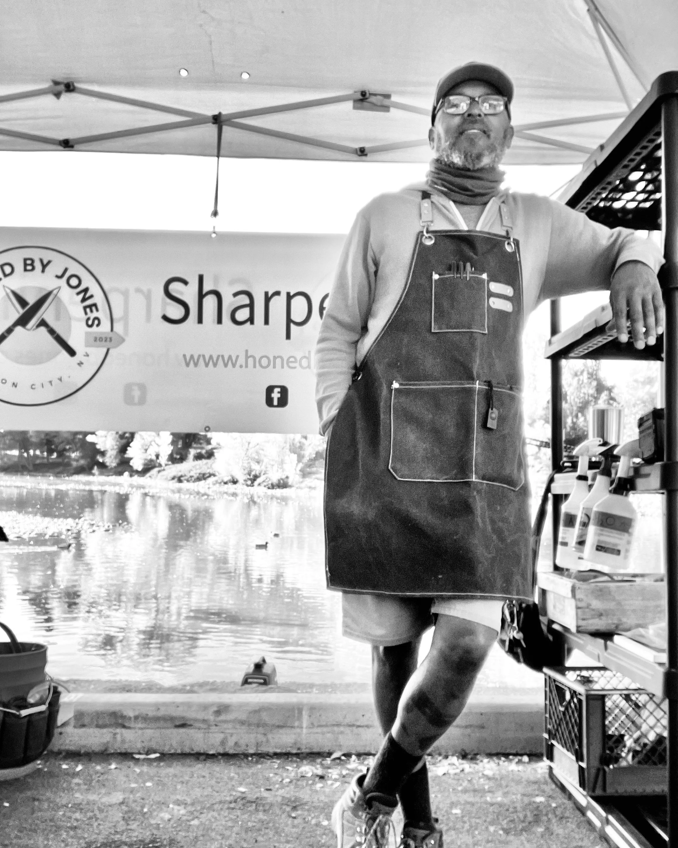 A person standing inside a tented area, wearing glasses, a cap, a neck gaiter, and an apron, with a paddleboard and water in the background.