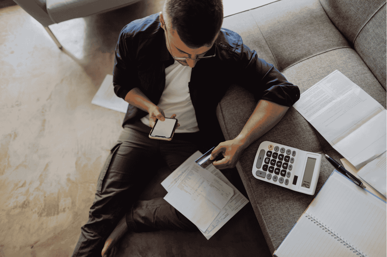 A young adult sits on the floor with a stack of utility bills on their lap. They are holding a cell phone in one hand and a credit card in the other. Surrounding them is a calculator, pen, notebooks, and more bills.