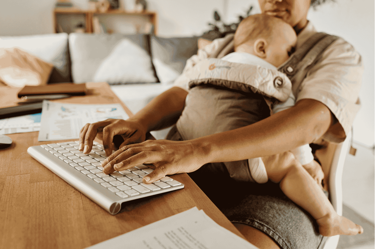 Image of a mother typing on a computer keyboard while holding a baby. In the background: utility bills and a check book.