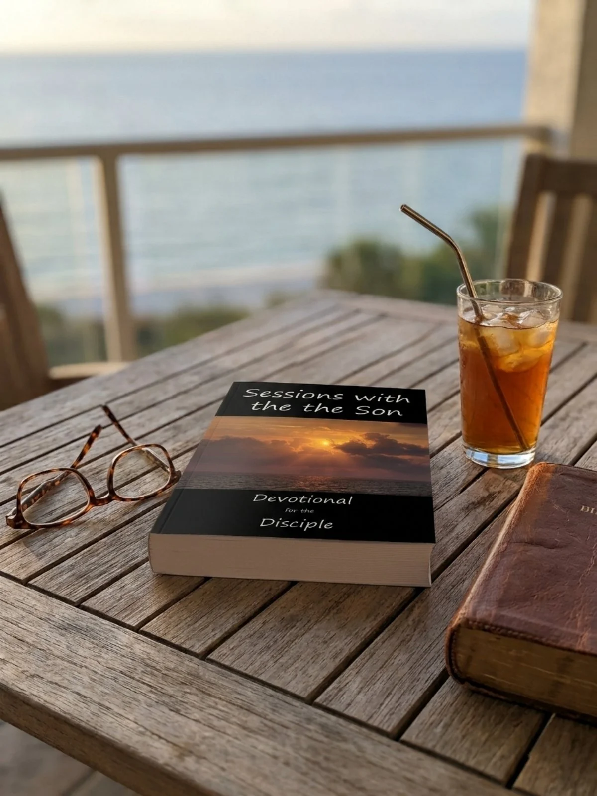 A wooden table with glasses of iced tea, a book titled 'Sessions with the Son: Devotional for the Disciple', a pair of reading glasses, and a leather-bound journal, with a blurred view of the ocean in the background.