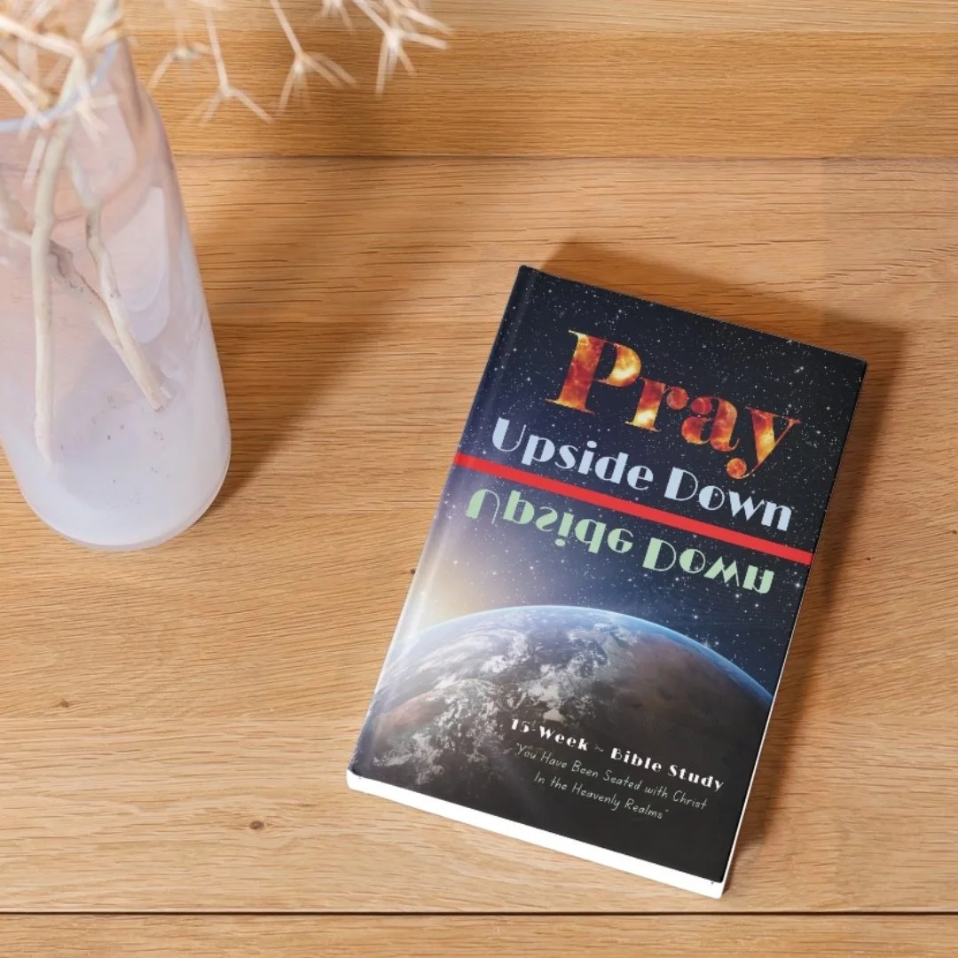 A Bible titled 'Pray Upside Down' resting on a wooden surface next to a white vase with dried plants.