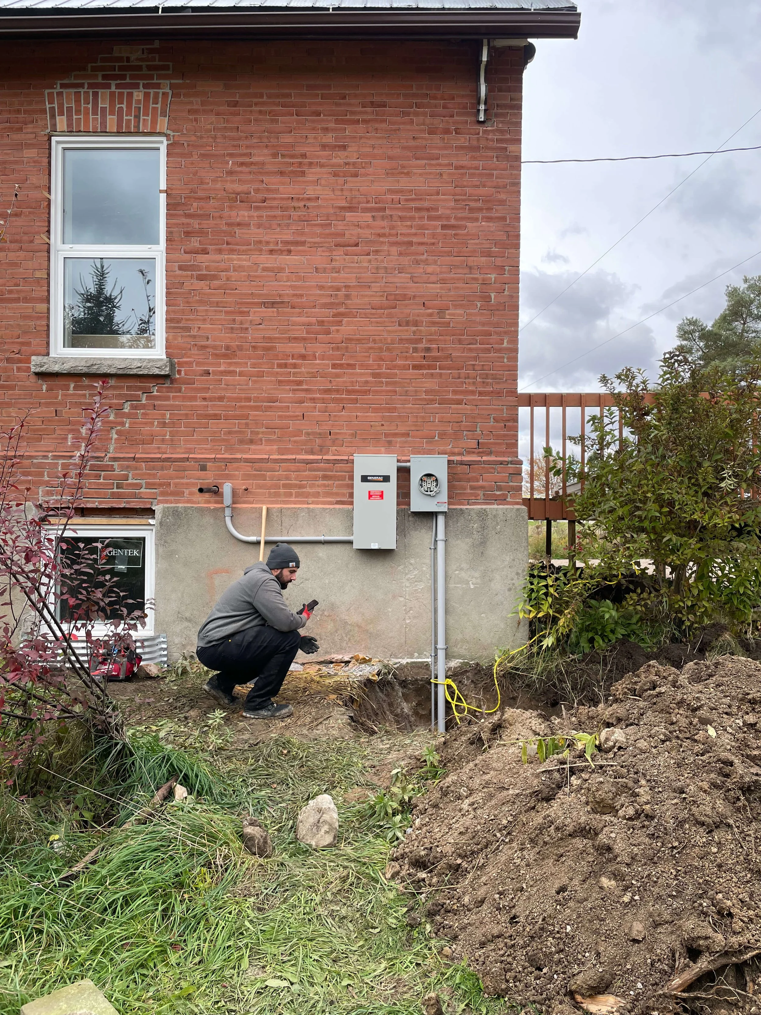 A person crouching next to an excavation site outside of a brick house, working on utility connections with electrical panels and wiring visible on the building exterior, cloudy sky overhead.