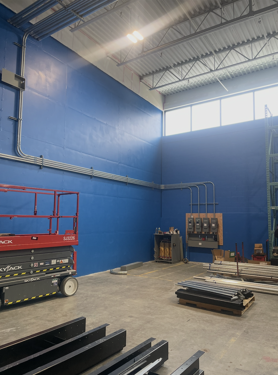 Empty industrial warehouse with blue walls, large windows, and various construction materials and equipment on the concrete floor.