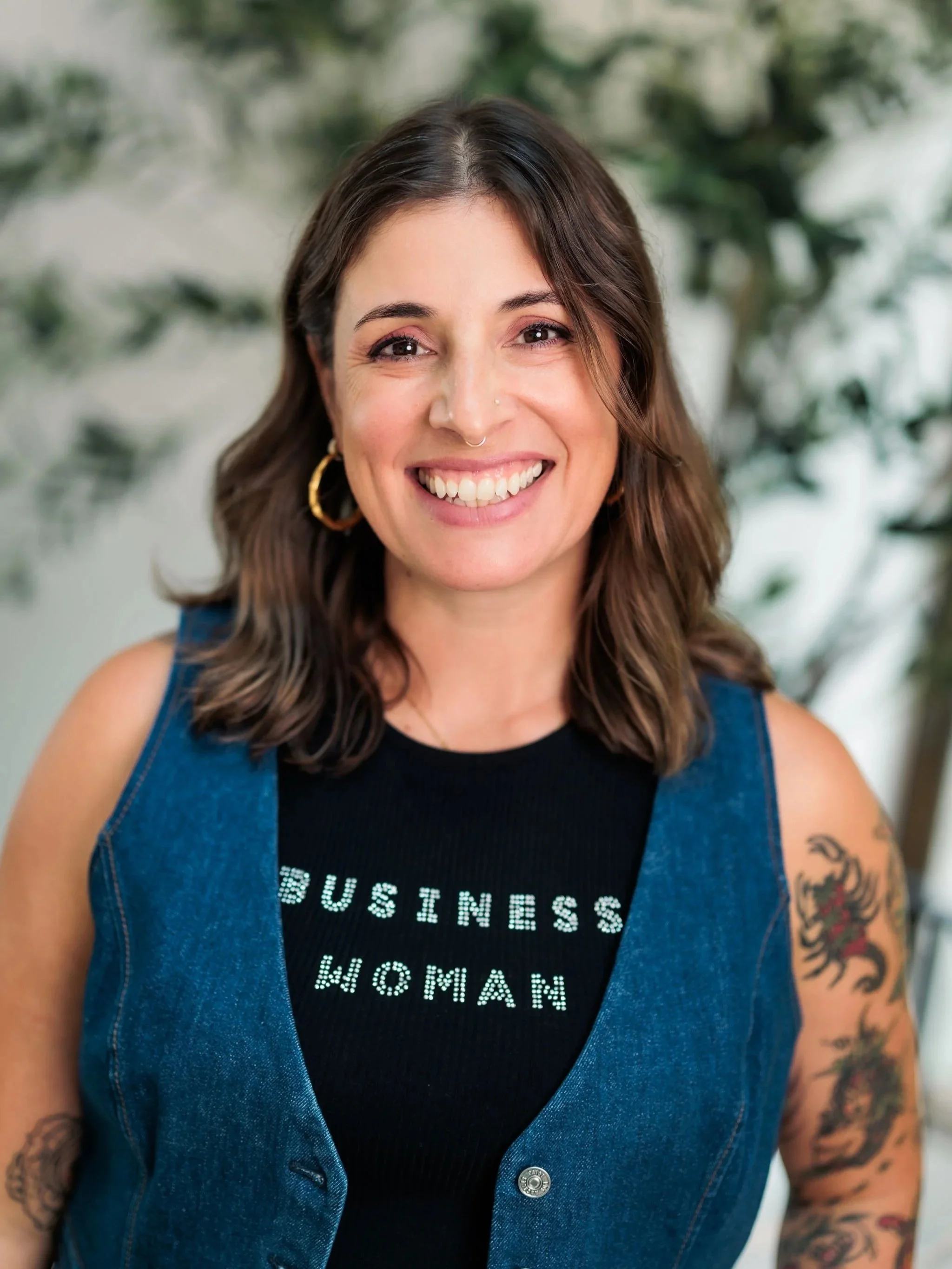 Close-up of a smiling woman with brown hair, tattoos on her arms, wearing a black T-shirt with white text and a sleeveless denim jacket, set against a blurred green background.