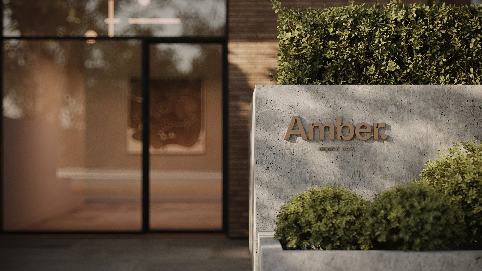 Concrete sign with the word 'Amber.' and smaller text 'Herne Bay,' surrounded by greenery, next to a building with glass and brick walls.
