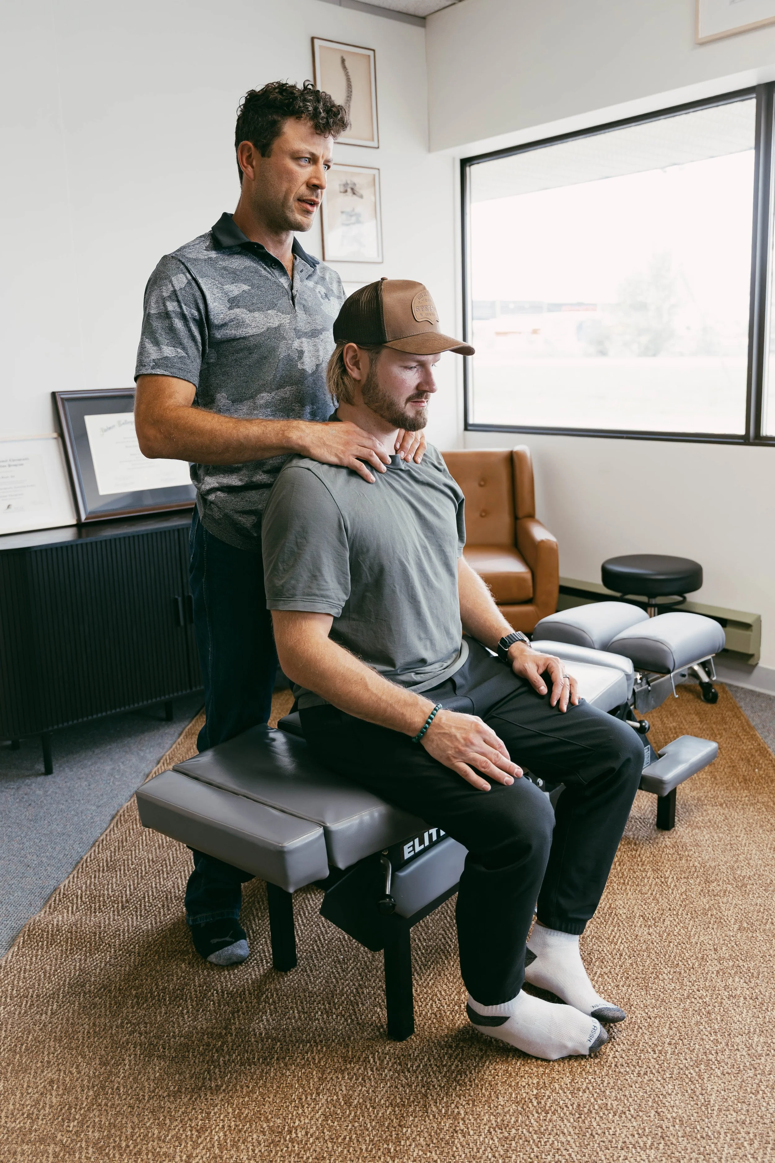 A chiropractor is performing an examination or treatment on a male patient who is seated on an examination table in a medical office. The chiropractor is standing behind the patient, supporting his neck with his hands, while the patient looks down with eyes closed. The room is well-lit with a large window, and framed anatomical artwork hangs on the wall.